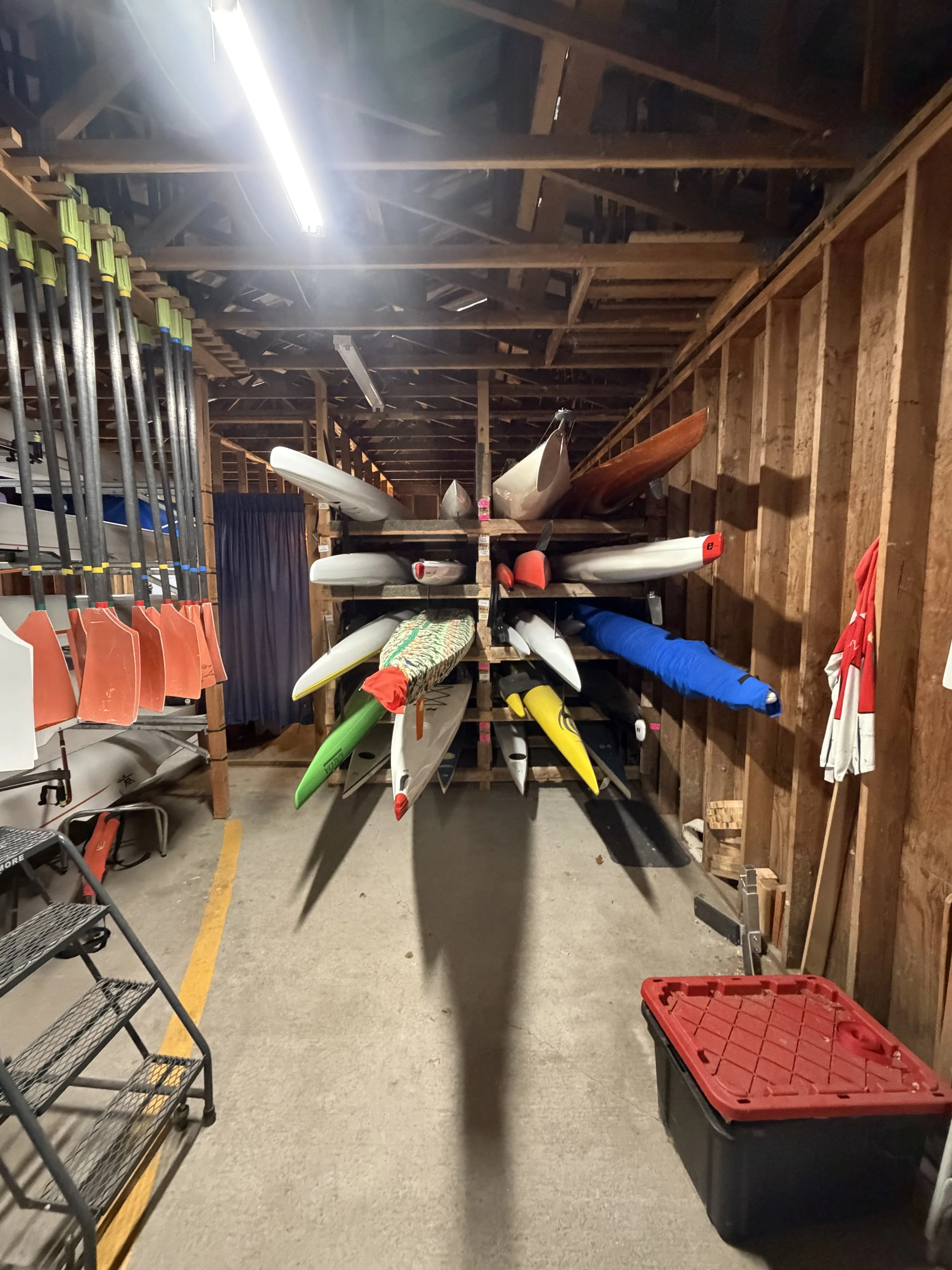 A storage room with boats, paddles, and sports equipment organized on shelves and against the walls, with a concrete floor and exposed wooden ceiling beams.