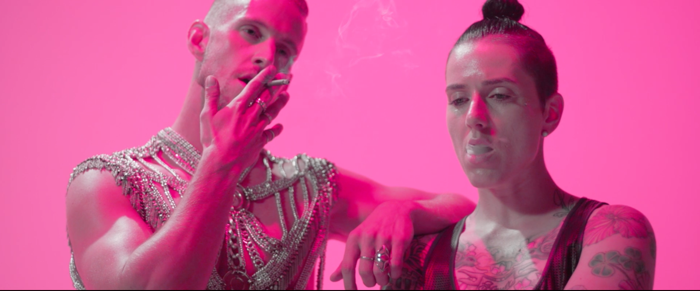 Two tattooed women with tattoos on their shoulders, one with hair in a bun, standing against a pink background. The woman on the left, wearing a pearl-studded outfit, is smoking a cigarette while the woman on the right is looking down.