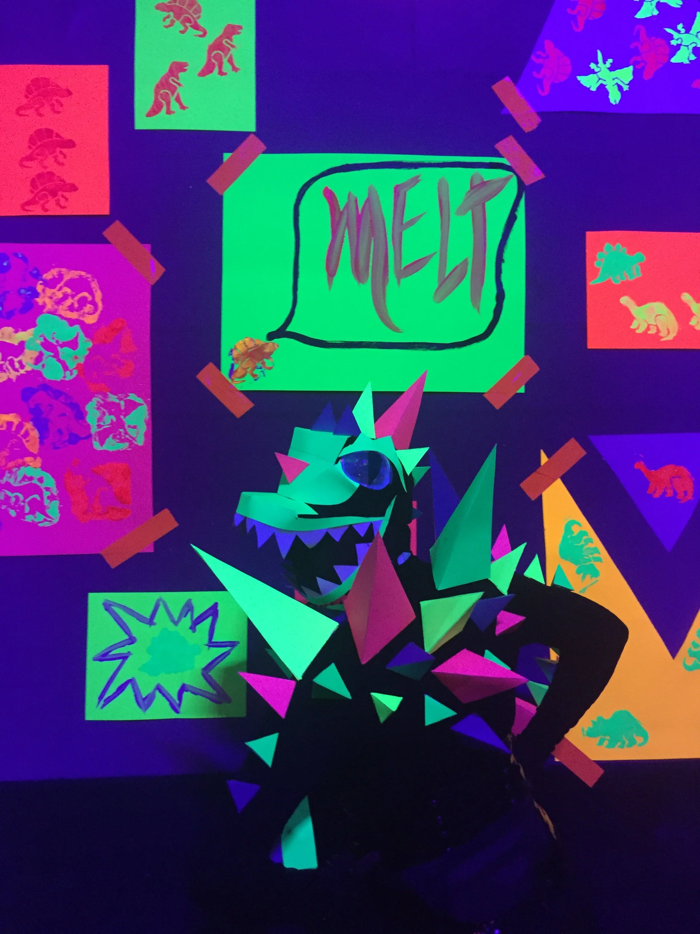 A colorful, abstract display with neon paper dinosaurs, stars, and chaotic shapes under blacklight, with the word 'MET' written on paper in the center.