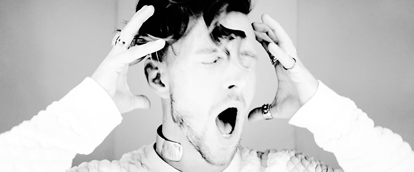 A young man with dark hair and facial hair, eyes closed, mouth open as if yawning or shouting, with hands on his head, wearing rings and a white sweater, in a close-up black-and-white photo.