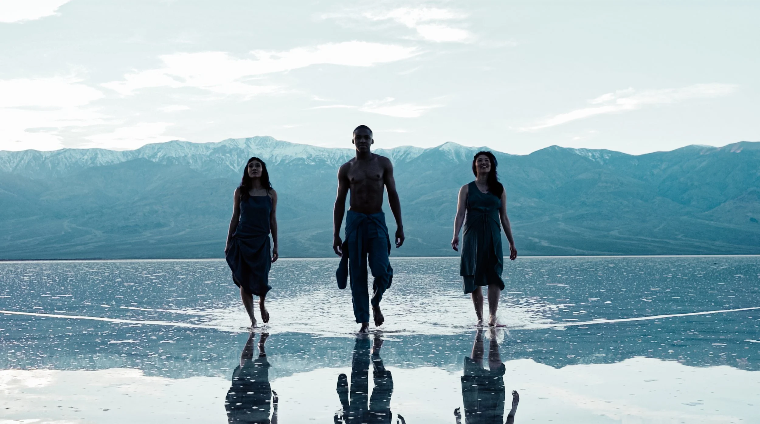 KINRGY performers walking through shallow water in a mountain landscape.