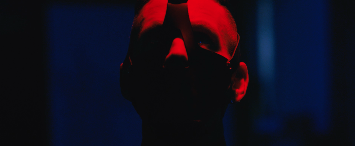 A person wearing sunglasses is illuminated with red and blue lighting in a dark setting.