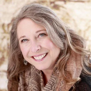 A woman with long, wavy gray hair smiling, wearing a brown patterned scarf and a black top.