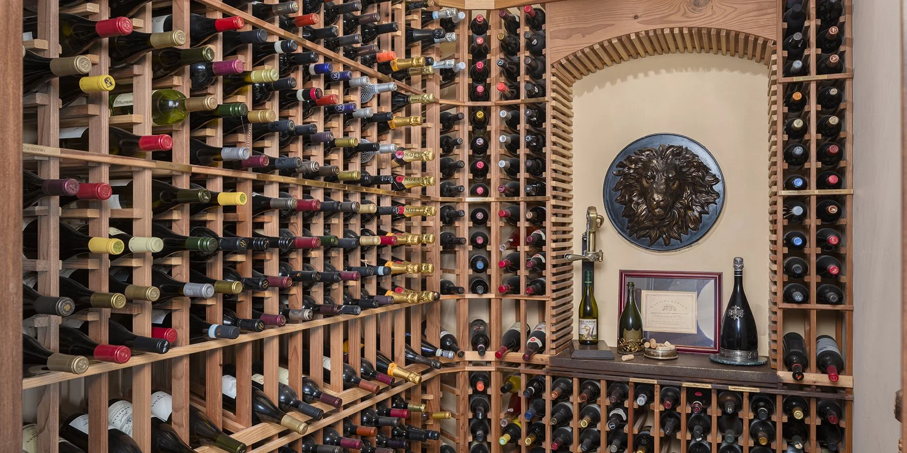 A wine cellar with wooden racks filled with various bottles of wine, a wall-mounted lion head sculpture, and framed certificates or documents.