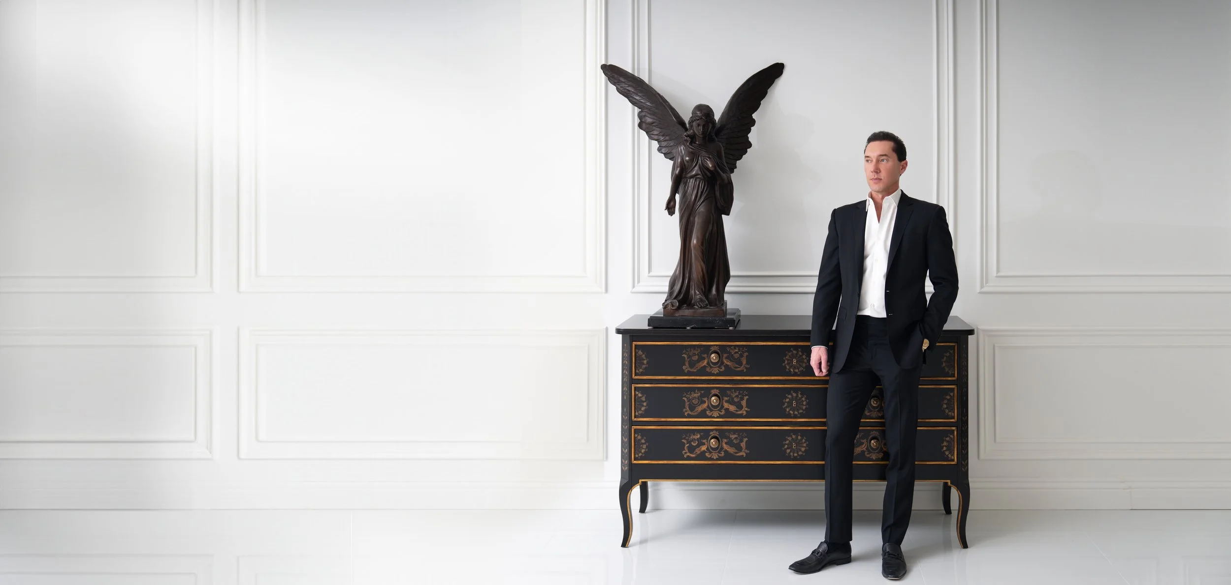 A man in a dark suit with a white shirt standing next to a black and gold ornate chest of drawers with a large dark sculpture of an angel with wings standing on top.