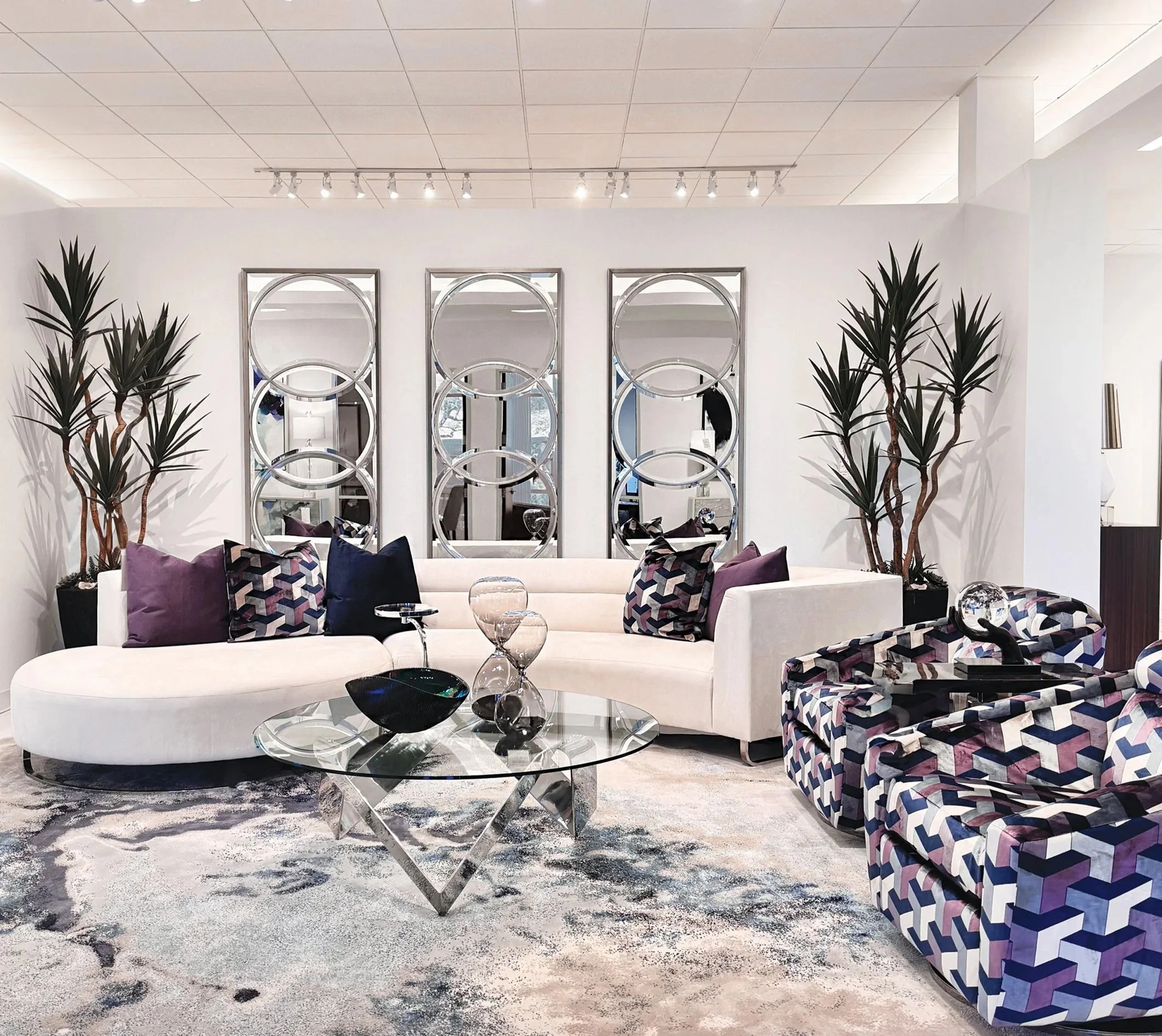 A modern living room with a white curved sofa adorned with purple and patterned pillows. There are two matching patterned armchairs, a glass coffee table with decorative glass vessels, and two tall potted plants flanking the sofa. Behind, three large circular mirrors hang on a white wall with ceiling track lighting above.