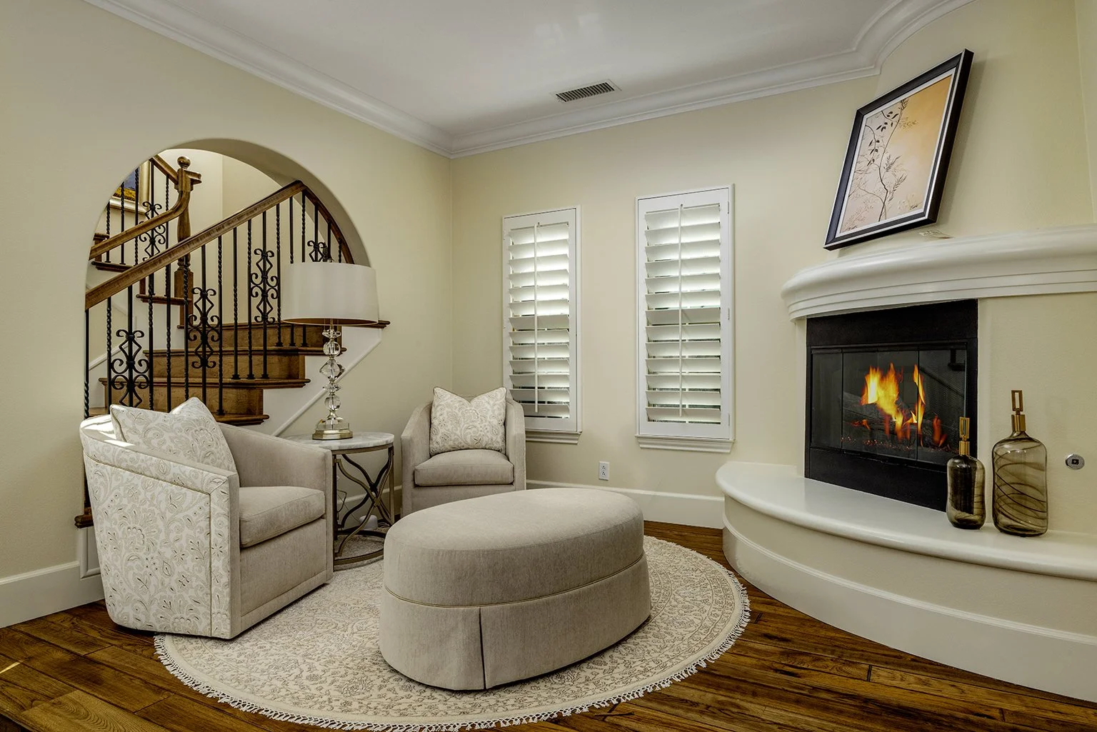 living room with fireplace, beige sofa, armchair, ottoman, wooden floors, two windows with plantation shutters, framed artwork above fireplace, decorative vases, and staircase with wooden railing and wrought iron balusters.