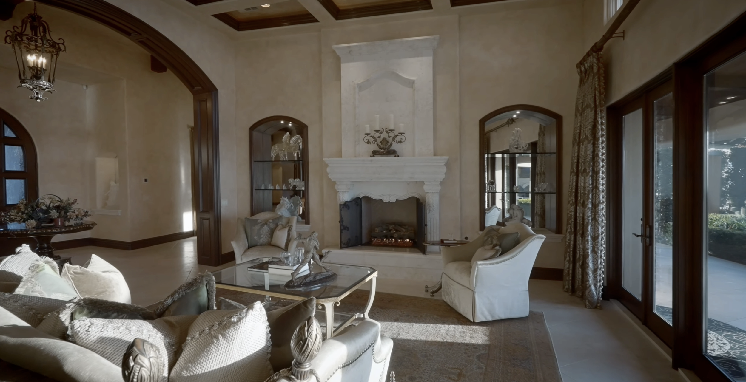 Elegant living room with cream-colored furniture, a large white fireplace, glass display cabinets, and large windows with patterned curtains, allowing natural light to fill the space.