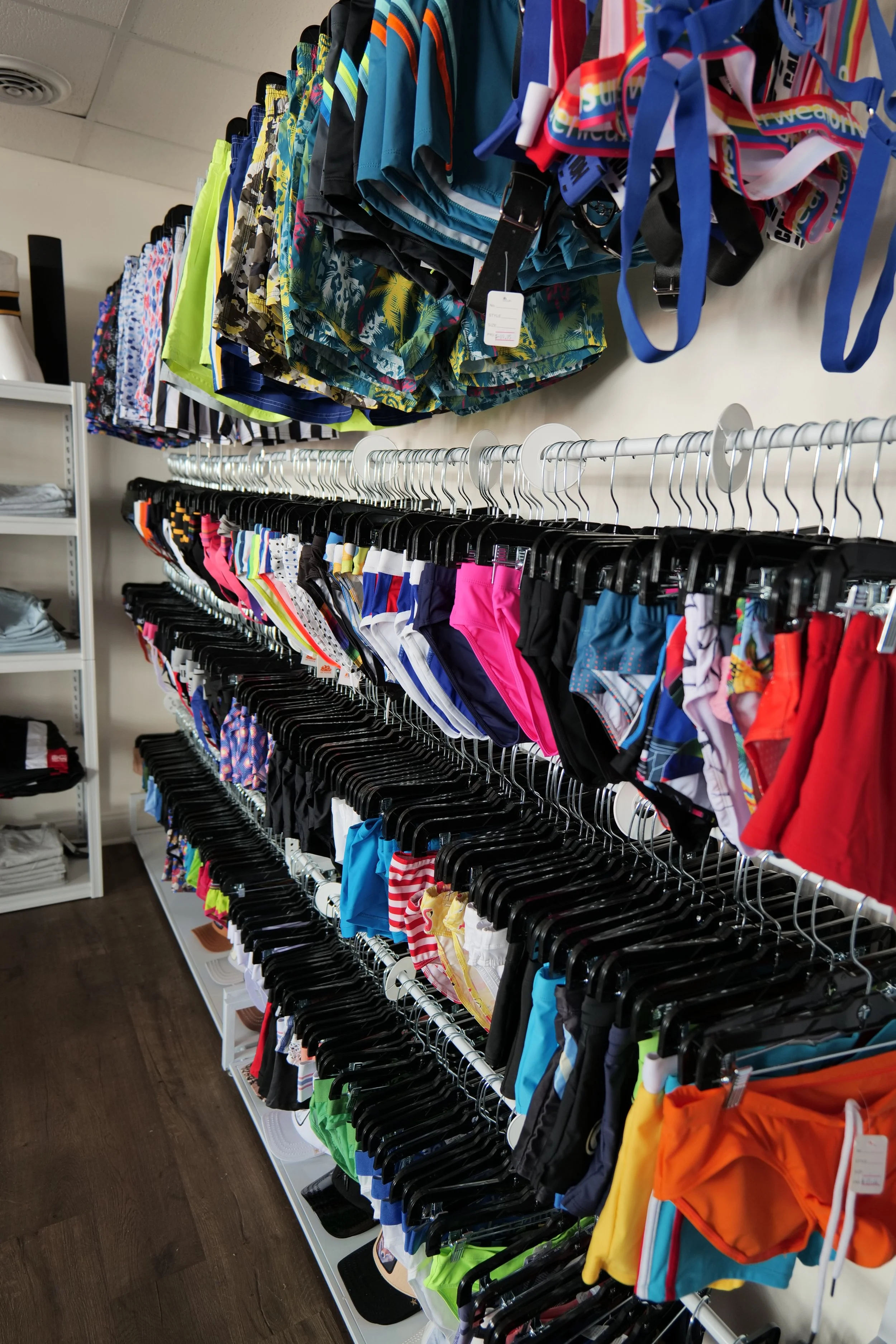 Close-up of multiple rows of colorful men's and women's swimwear hanging on metal racks in a retail store, with additional swimwear on the upper shelf and shelves of folded clothing on the left side.