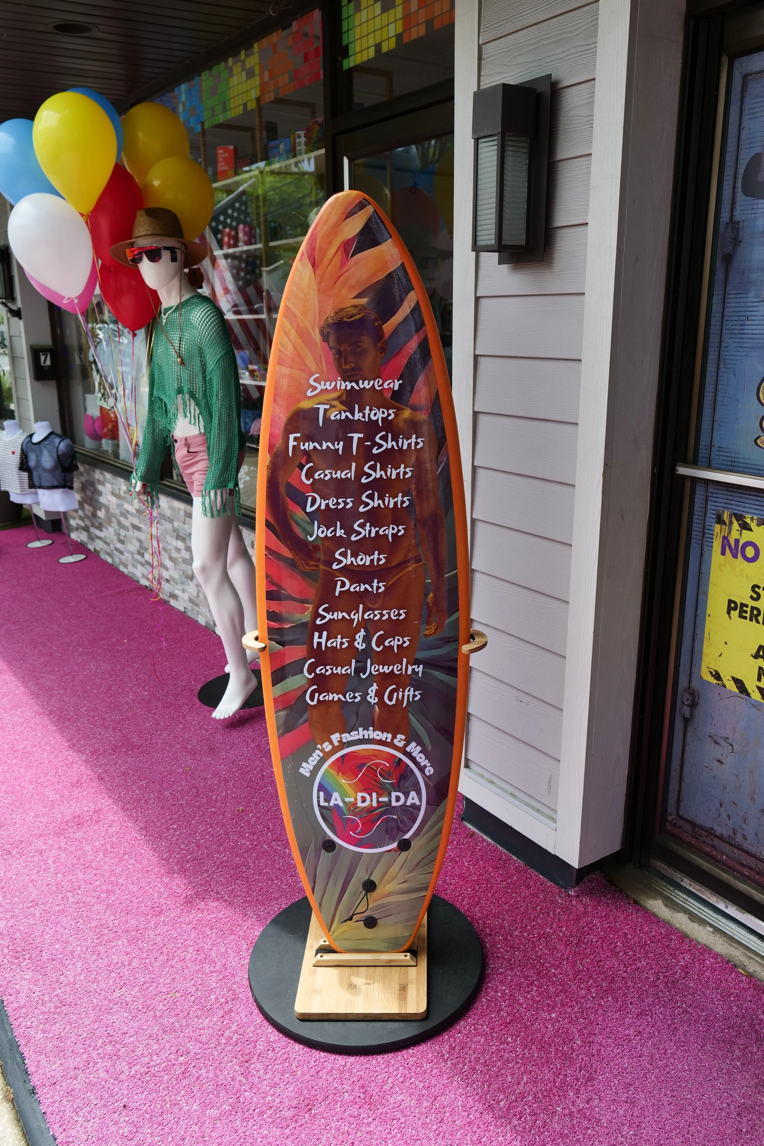 A surfboard-shaped sign listing products such as swimwear, tank tops, funny t-shirts, casual shirts, dress shirts, jock straps, shorts, pants, sunglasses, hats & caps, casual jewelry, games, and gifts outside a clothing store with mannequins dressed 