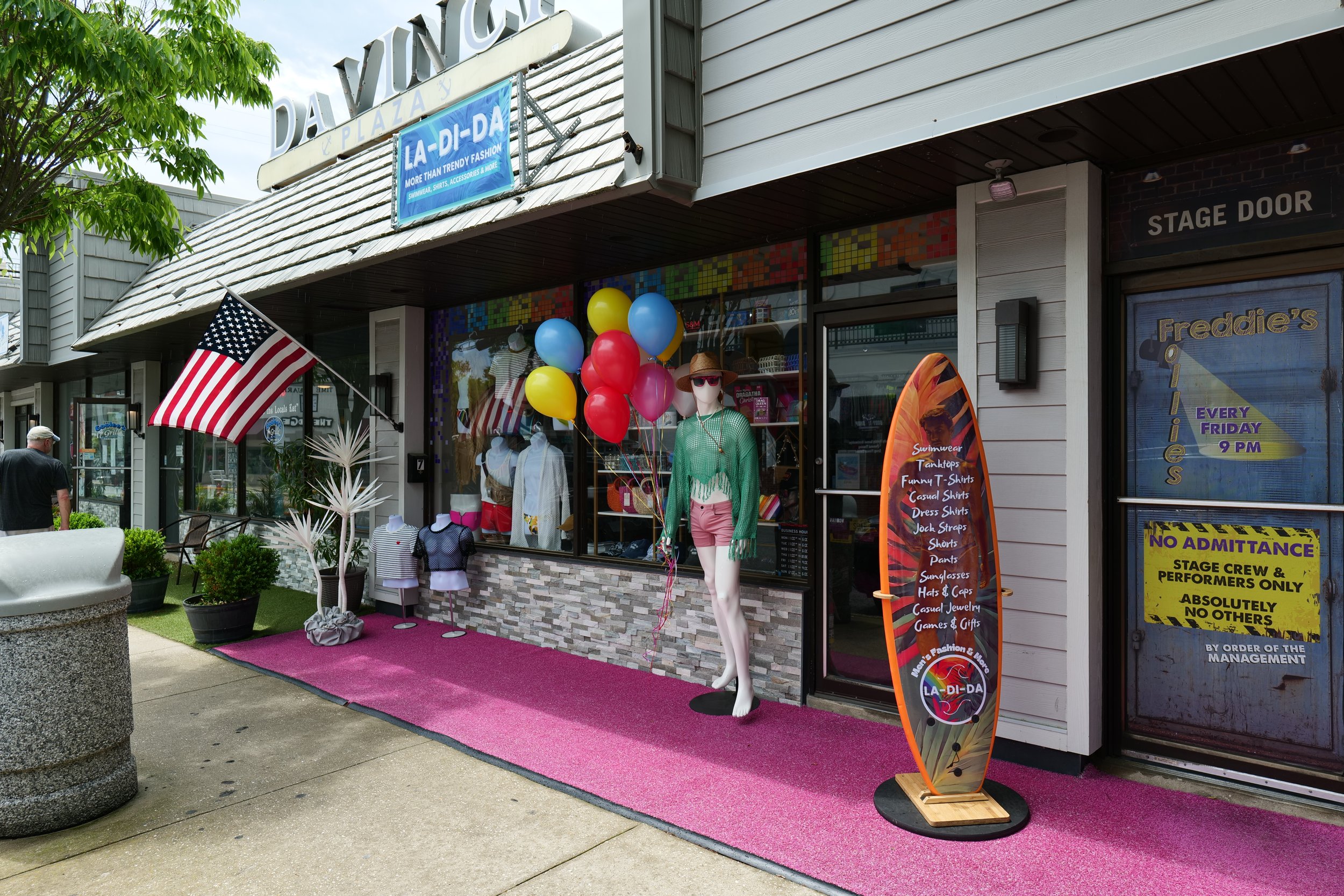 Storefront display with pink carpet, mannequin in green top and pink shorts, colorful balloons, American flag, and surfboard sign for La-Di-Da store.
