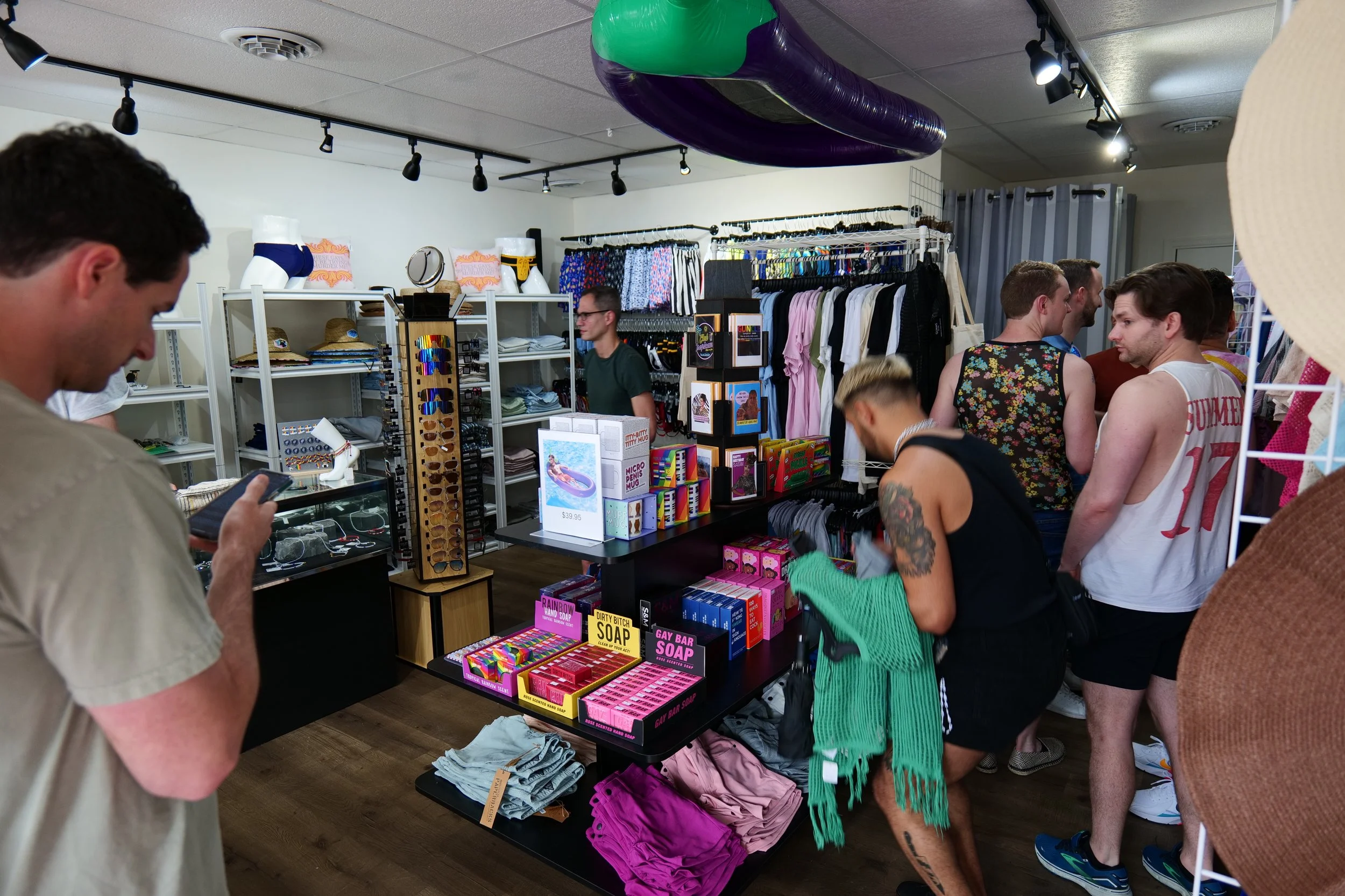 Inside a retail store with shelves of clothing, accessories, and novelty soap, with several customers browsing and one person looking at their phone.