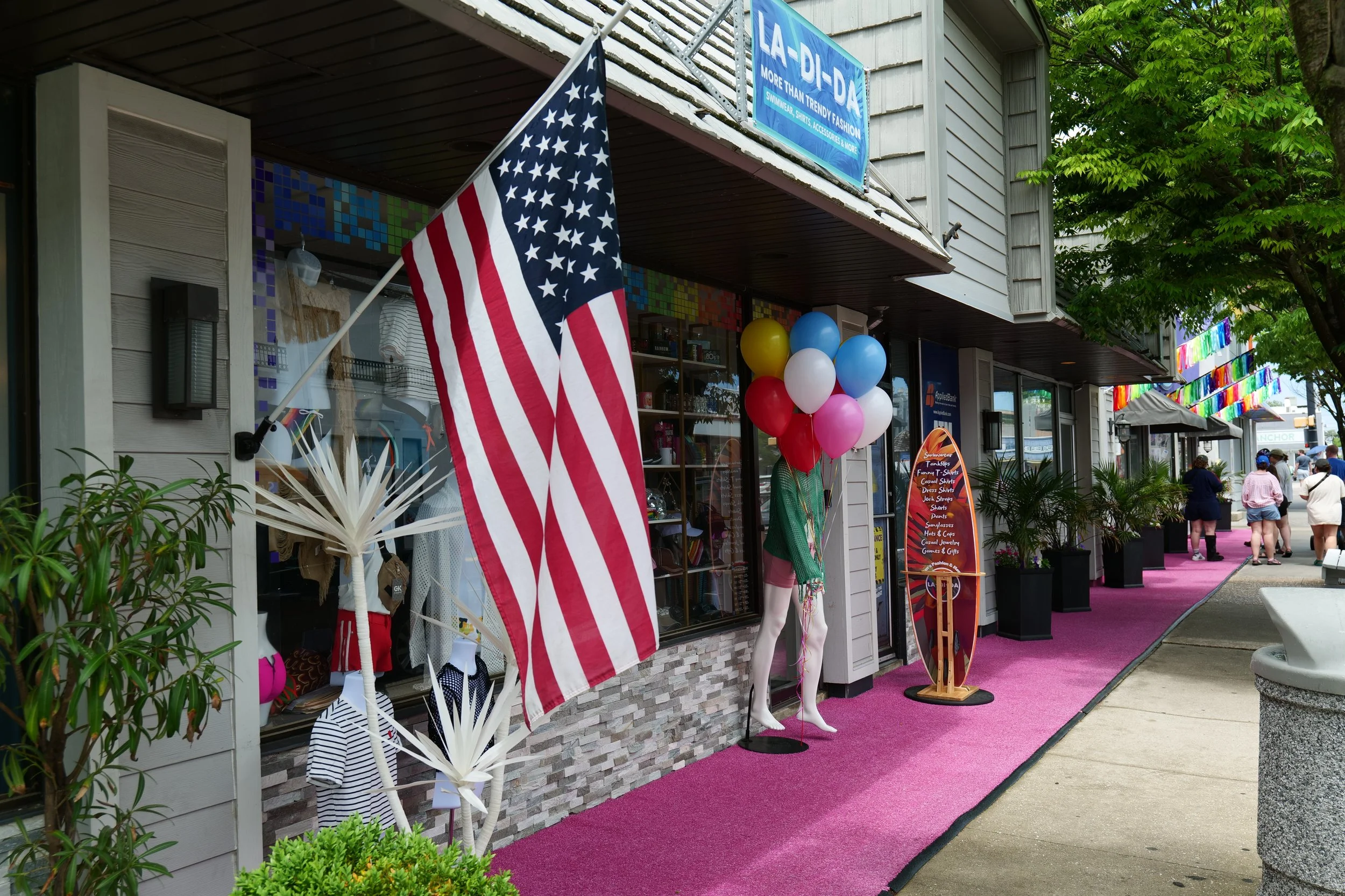 Street view of a boutique shop decorated with an American flag, colorful balloons, and a pink carpet, with a sign and people walking by.