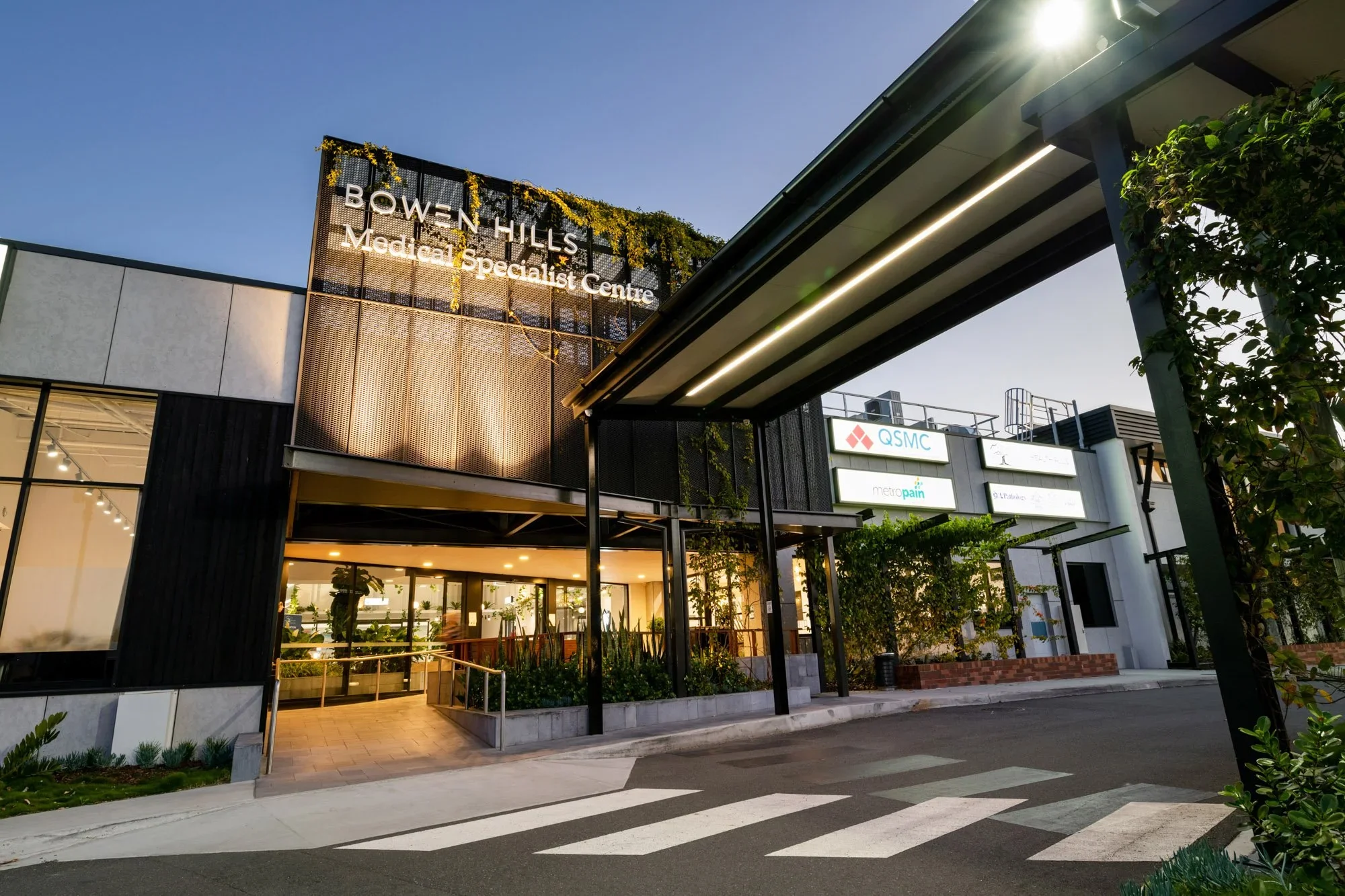 Exterior of Bowery Hills Medical Specialist Centre building with illuminated signage, entrance, and outdoor seating area under modern awning, with trees and clear sky in the background.