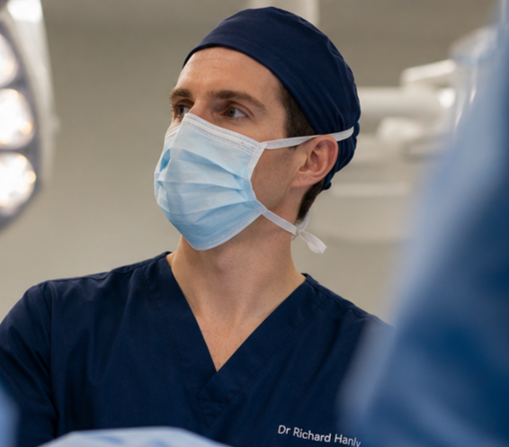 A male surgeon in scrubs, wearing a blue cap and face mask, in an operating room.