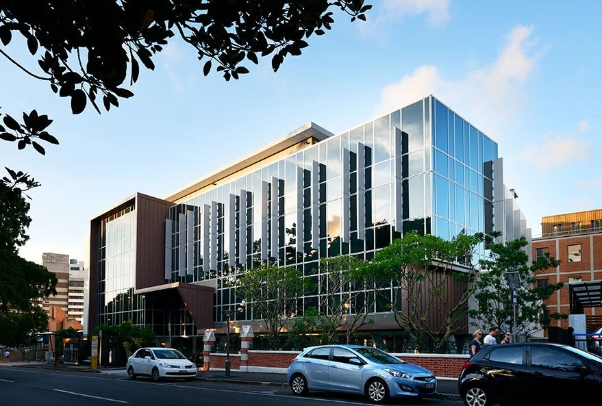 Modern multi-story glass building reflecting the sky, with cars parked in front and trees along the street.