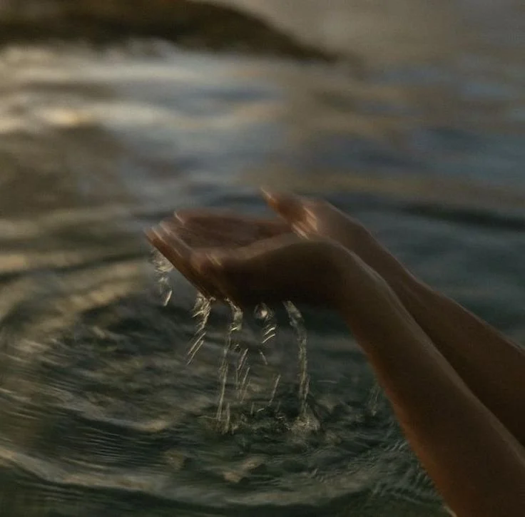 A person holds water in their hands over a body of water, with water dripping from their hands.