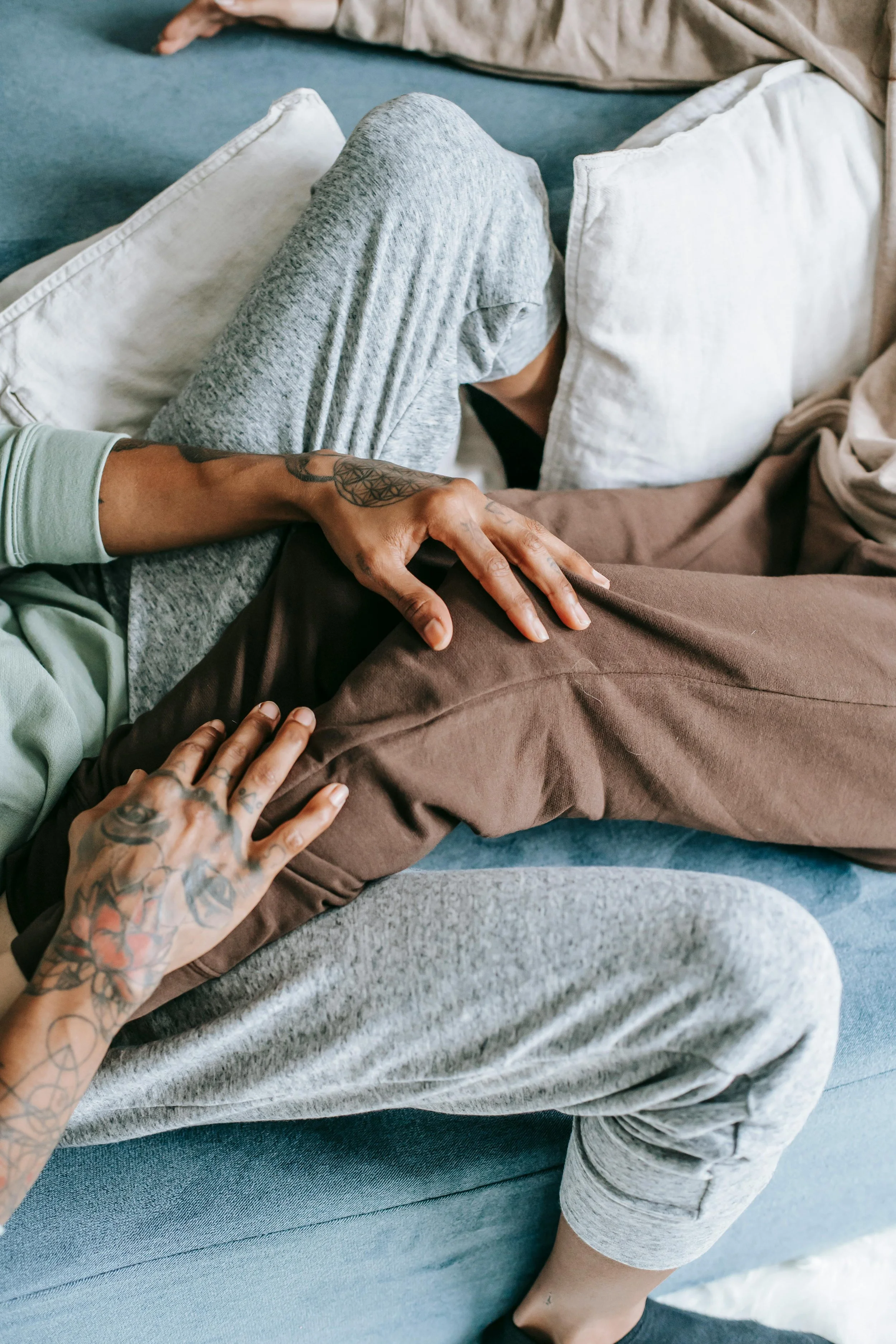 Person resting on a blue sofa with their head covered with pillows, while another person with tattoos holds their leg.
