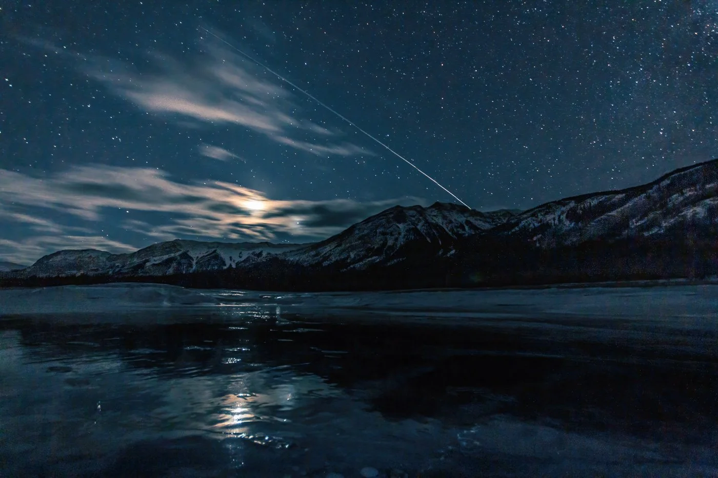 Star link satellite, stars, moon reflected on ice frozen lake Alberta Canada