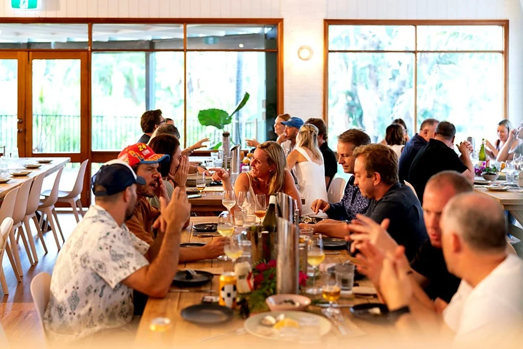 A group of people gathered around a long dining table in a bright, modern restaurant, engaged in conversation and enjoying drinks.