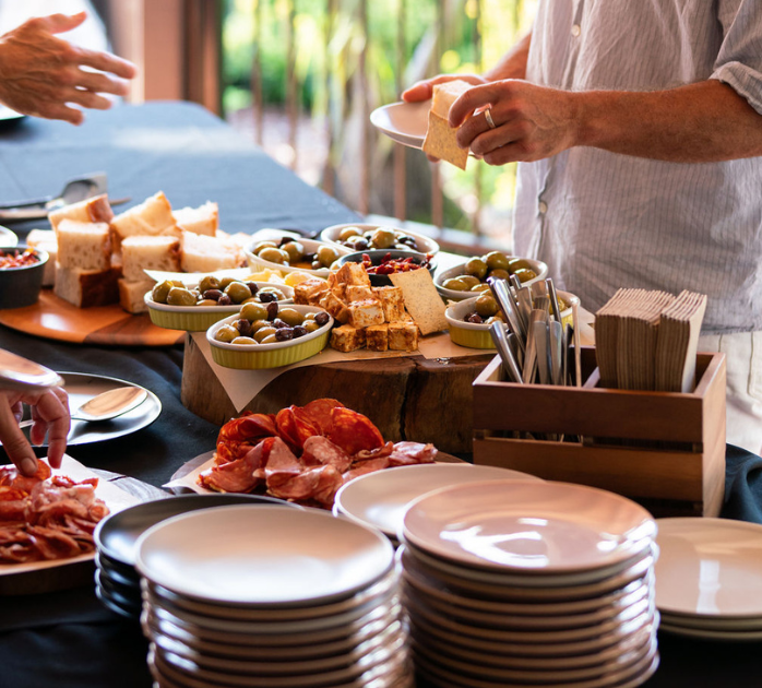 Charcuterie spread with bread, olives, and cheese, with a person serving from a plate, on a table set for a gathering.