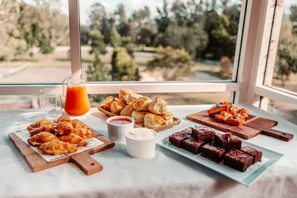 Assorted baked goods and breakfast items on a table near a window, including croissants, scones, brownies, and small bowls of sauces or dips, with a pitcher of orange juice and empty glasses.