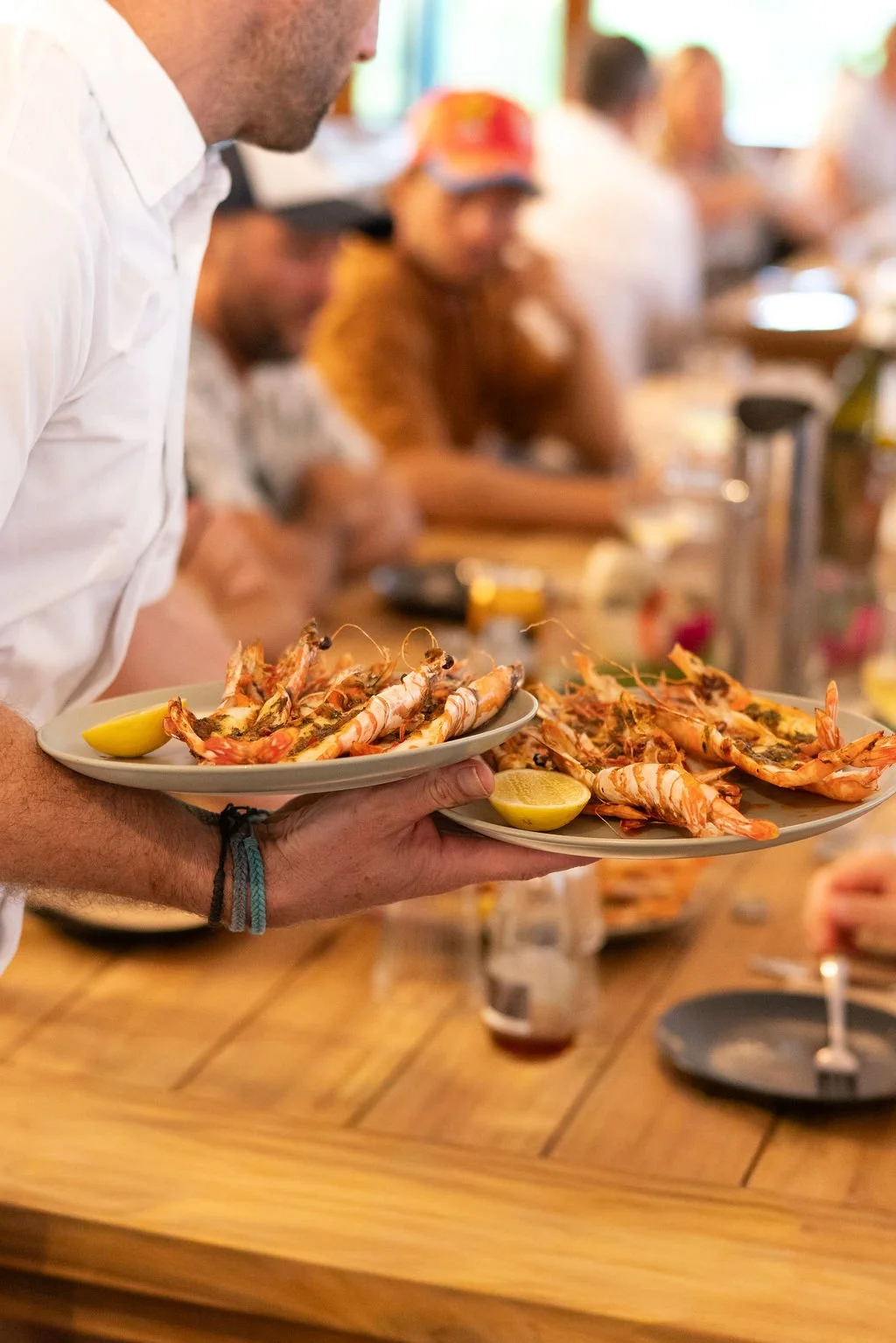 Person holding two plates of grilled shrimp with lemon wedges at a lively dining event.