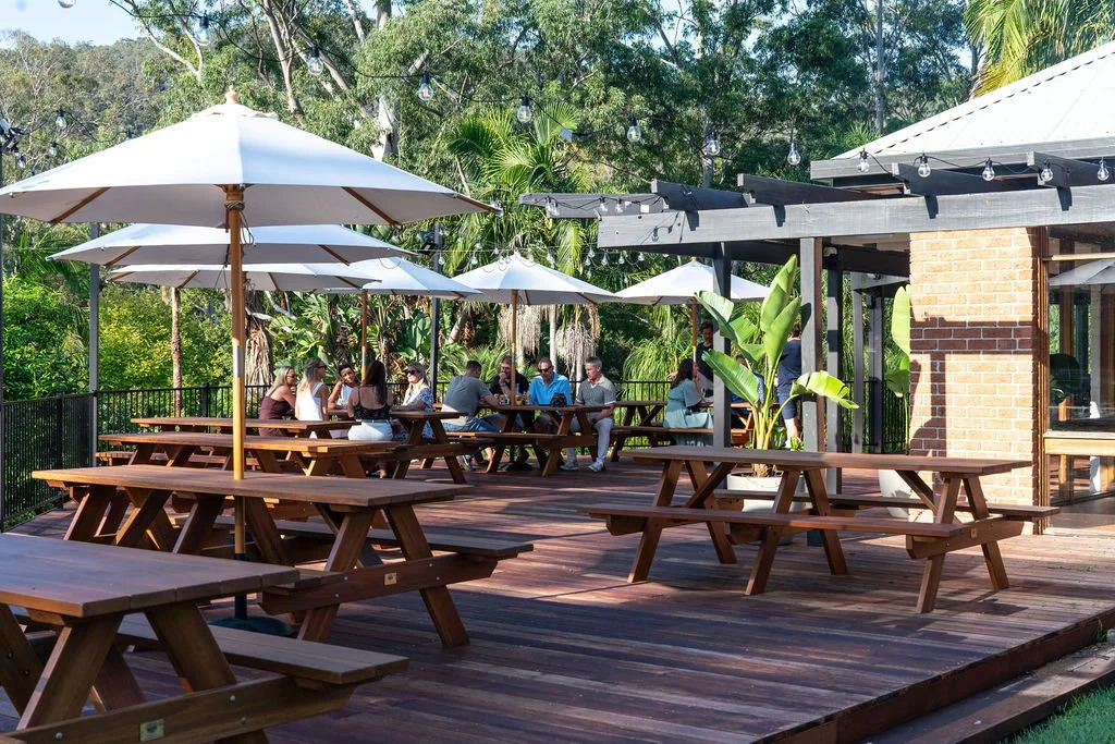 An outdoor patio with wooden picnic tables and white umbrellas, surrounded by lush green trees and string lights, with people sitting and socializing.