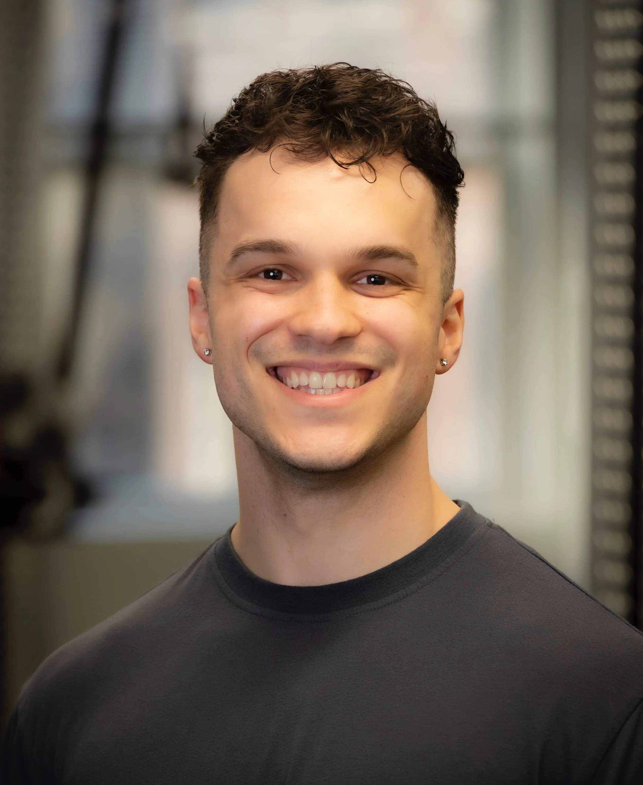 A young man with short, curly brown hair, earrings in both ears, smiling, wearing a black shirt, with a blurred background.