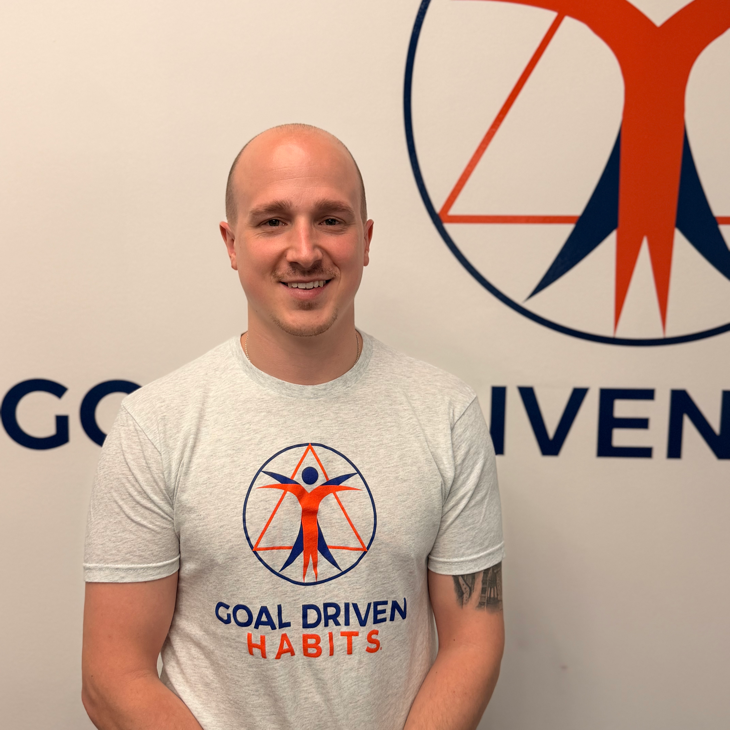 Coach Adam Fagan smiling in front of a wall with a logo and the text 'GOAL DRIVEN HABITS' on his gray T-shirt and the wall behind him.