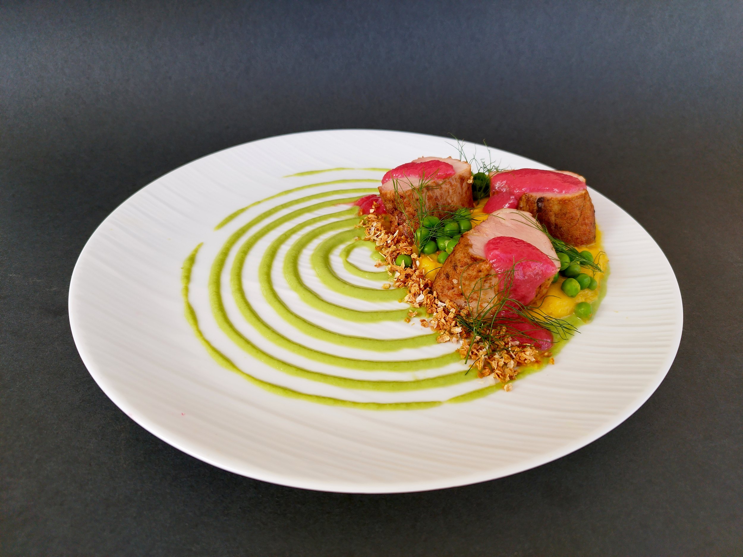 An artfully plated dish featuring three pieces of cooked meat, topped with pink sauce, with green peas, sprigs of dill, and a bed of toasted nuts or grains, all on a white plate with green sauce spirals on a black background.