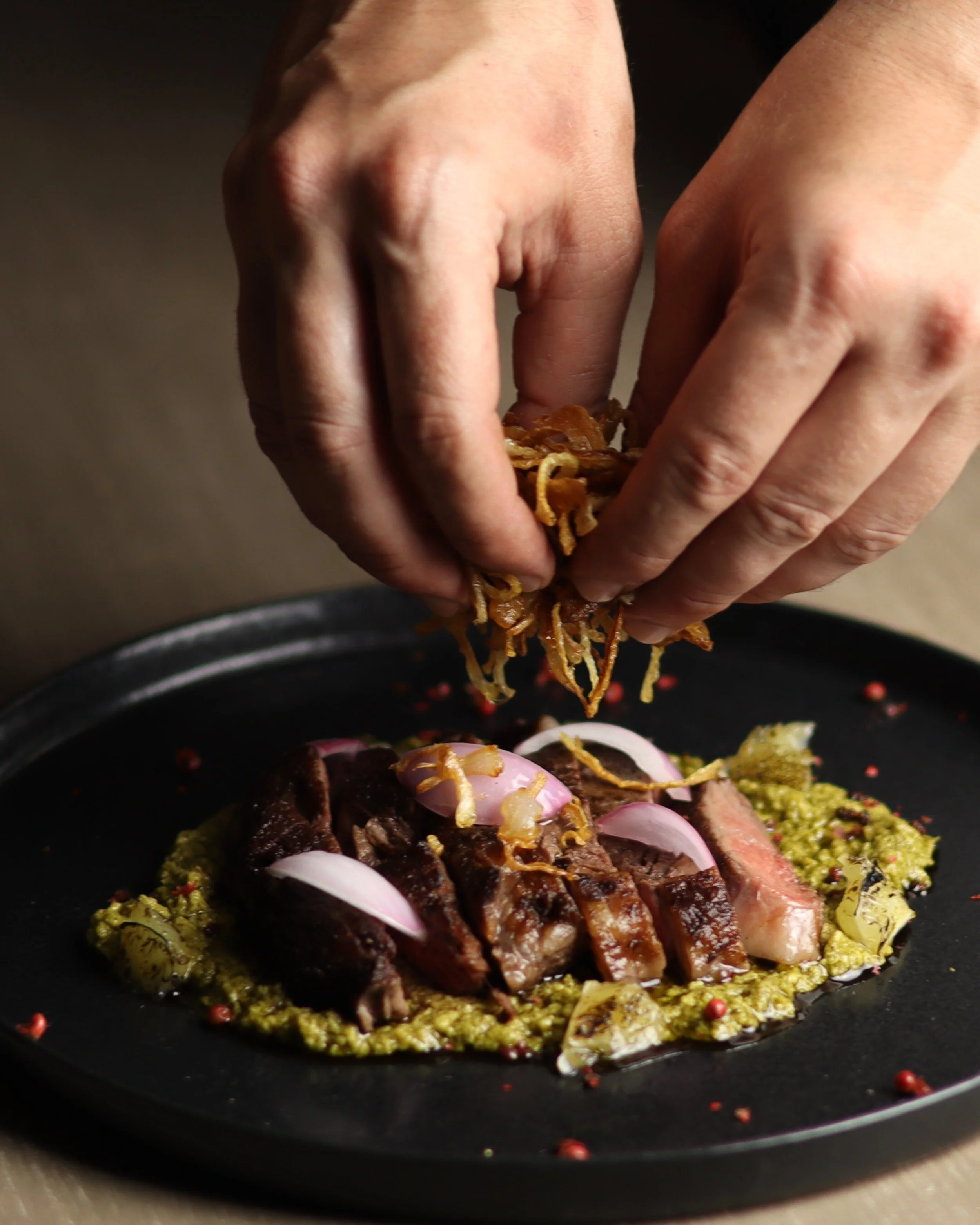 Close-up of hands garnishing a plate of cooked meat with fried onion strings, served with red onion slices, herbs, and sauces.