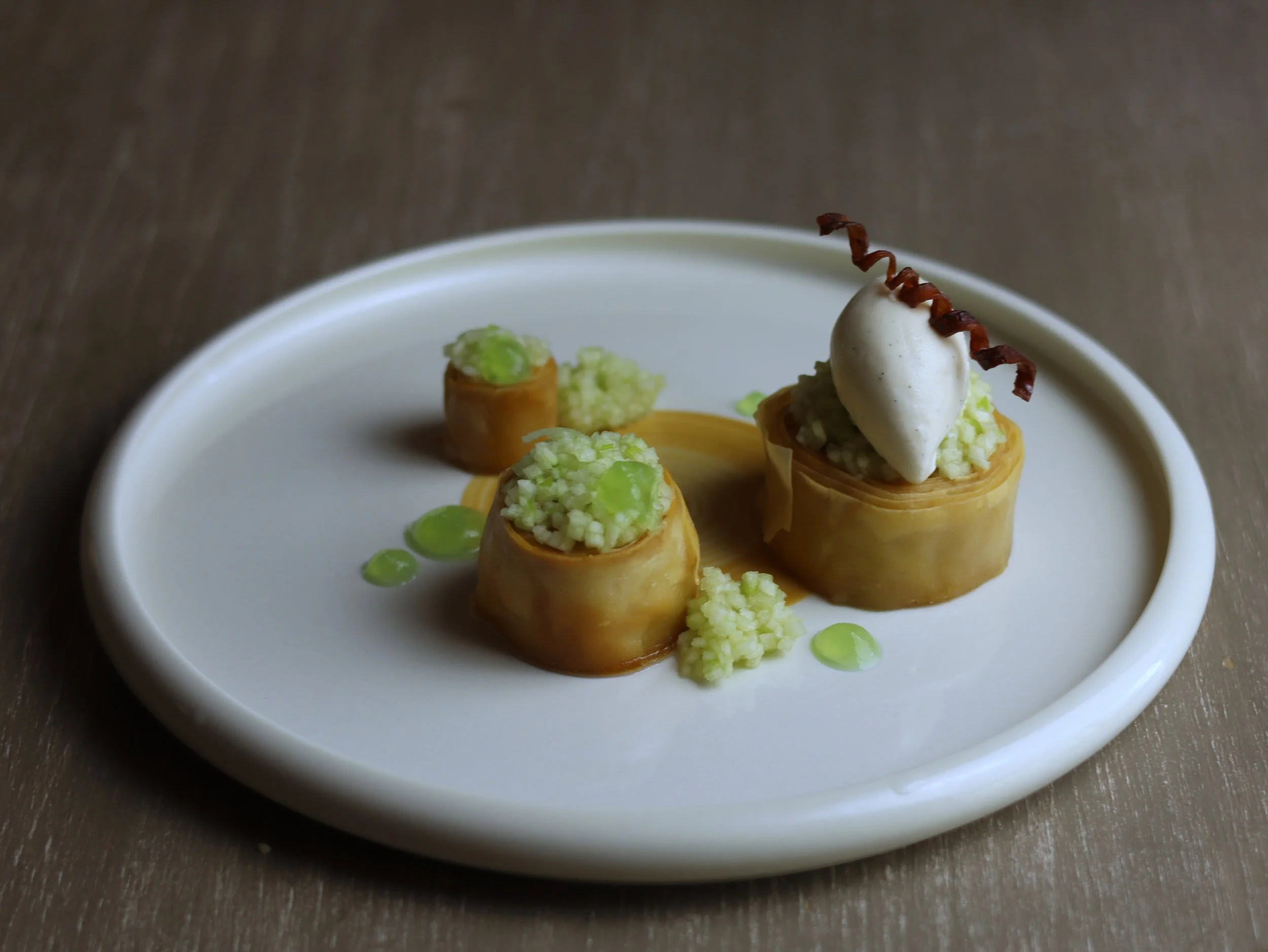 Dessert with small pastry shells filled with green cream, topped with white dollops and garnished with red spiral-shaped decoration, on a white plate with green sauce dots.