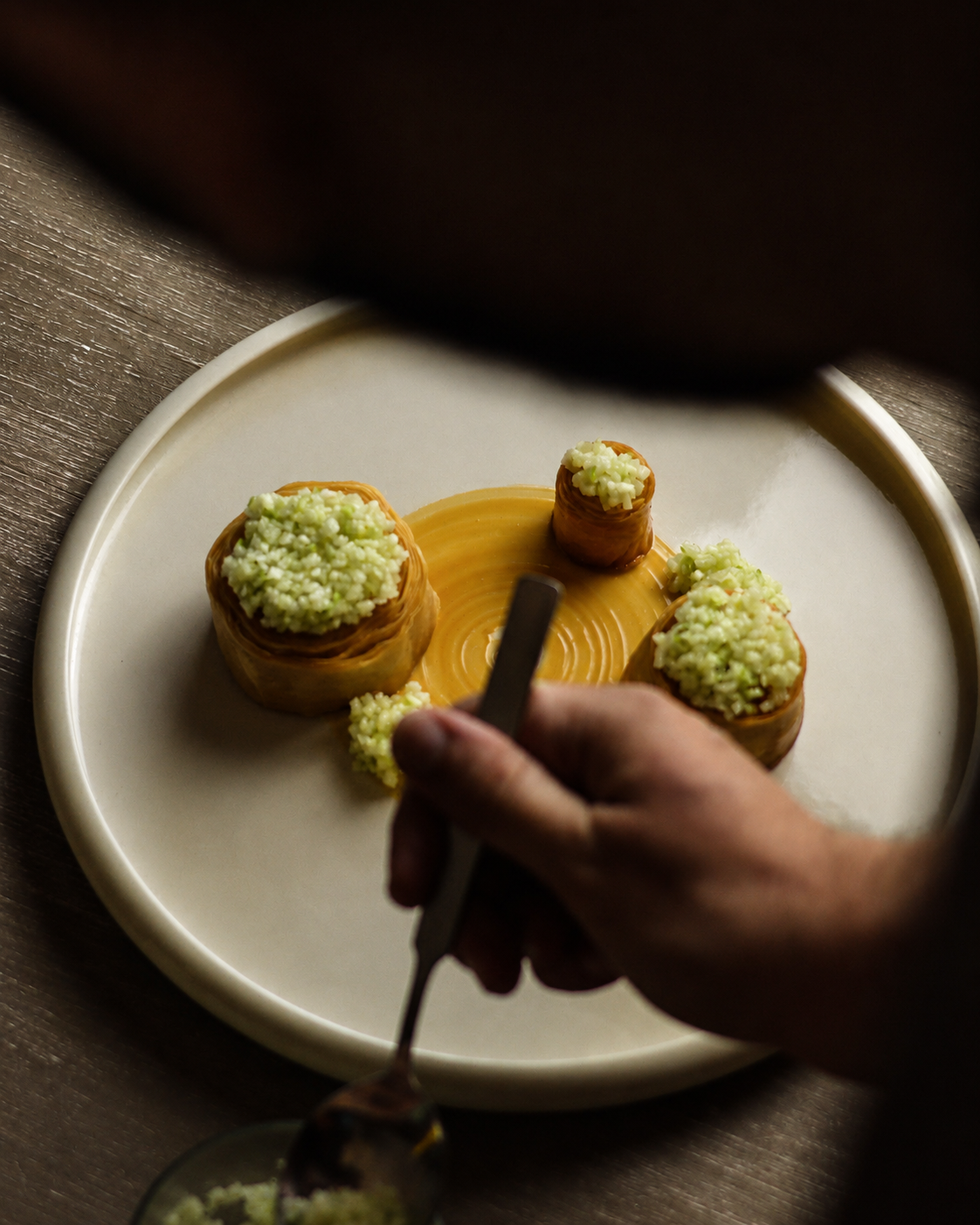 A chef's hand decorating small rolls of pastry filled with green stuffing on a white plate using tweezers.