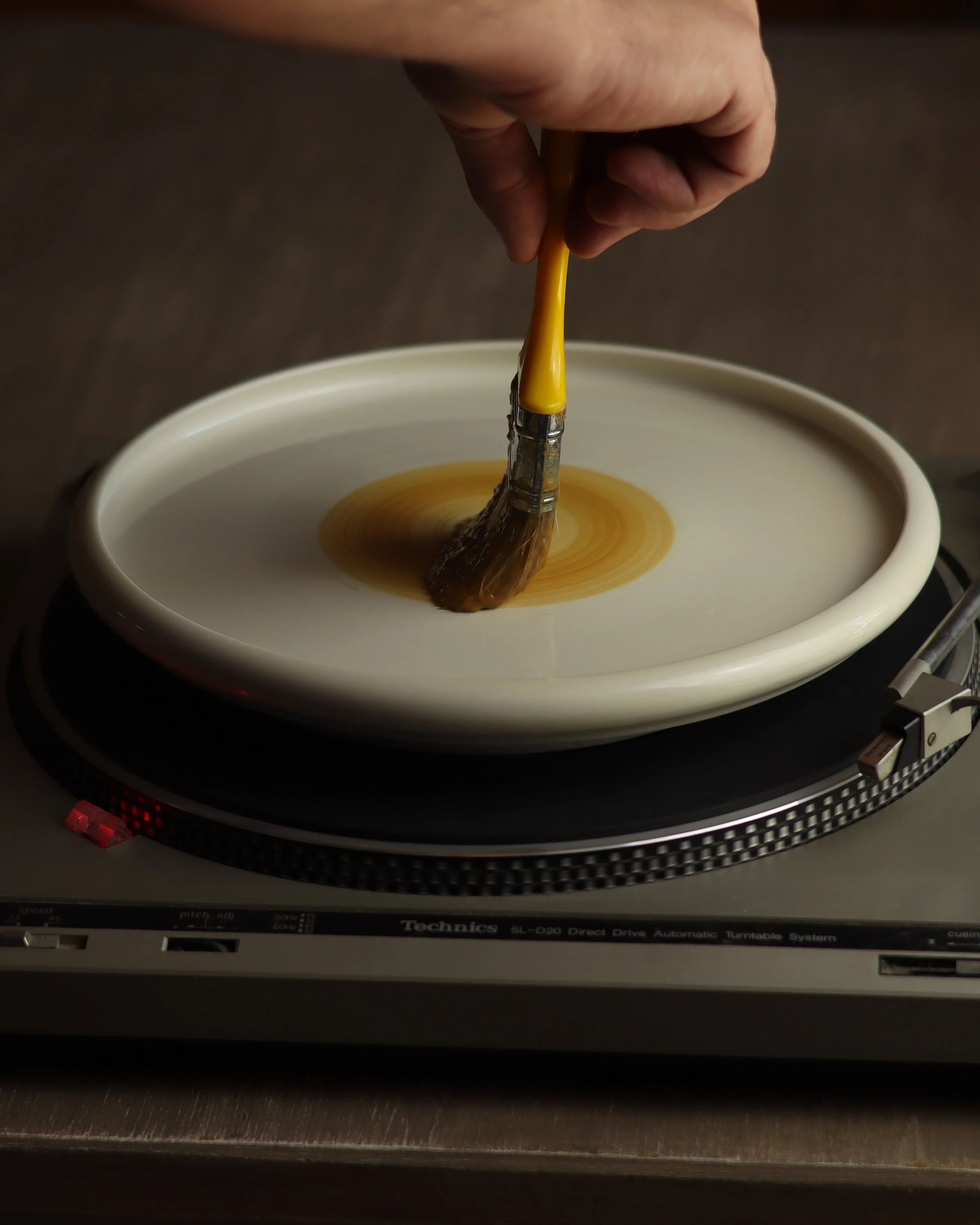 A hand is dipping a paintbrush into a small, white ceramic dish filled with a brownish liquid. The dish is placed on a turntable or record player. The background is dark and blurred.