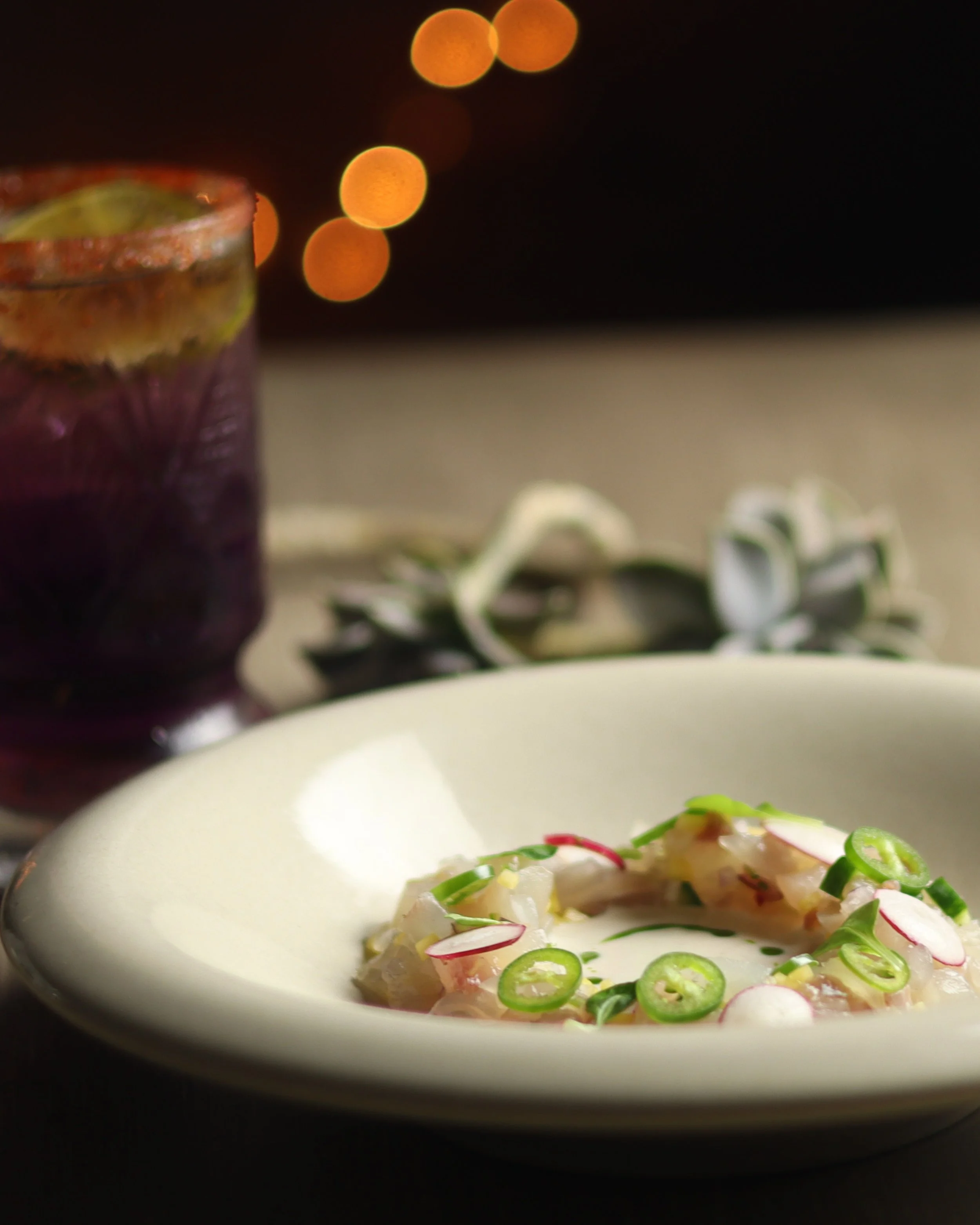 A white bowl filled with sashimi topped with sliced green onions and radish, with a blurred background featuring bokeh lights.