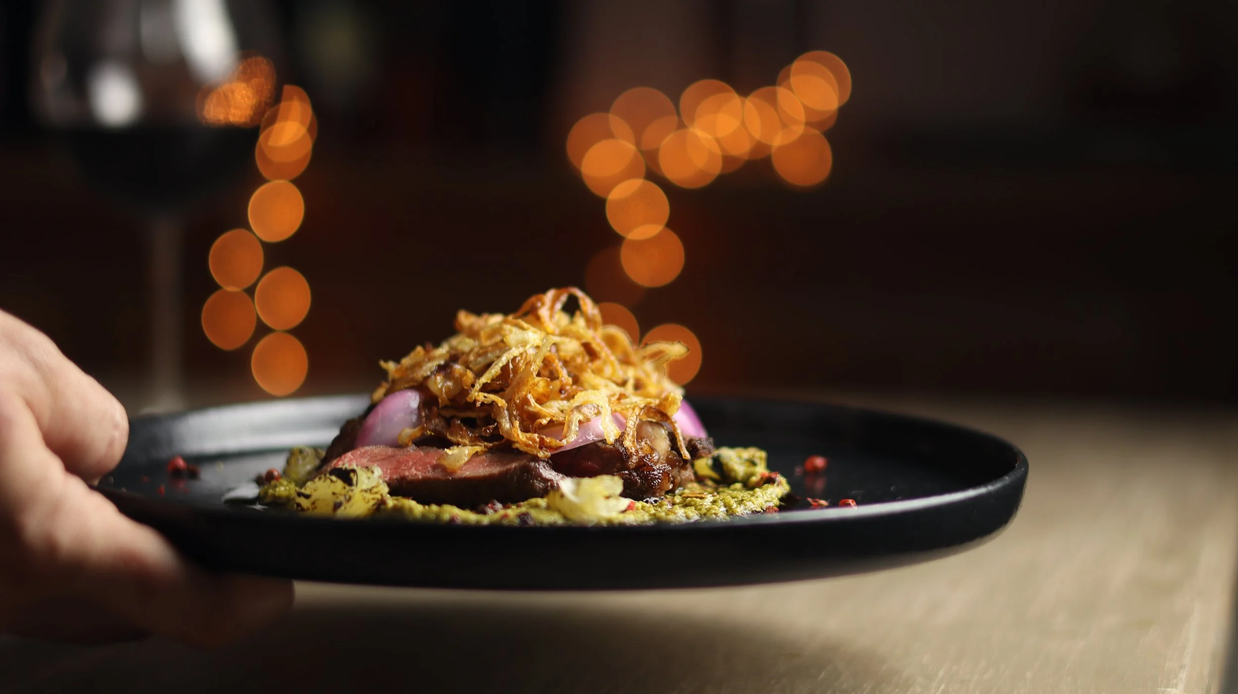 Close-up of a plate of gourmet food with a heap of crispy fried onions on top, fresh vegetables around, and a smoky, dark background with orange bokeh lights.