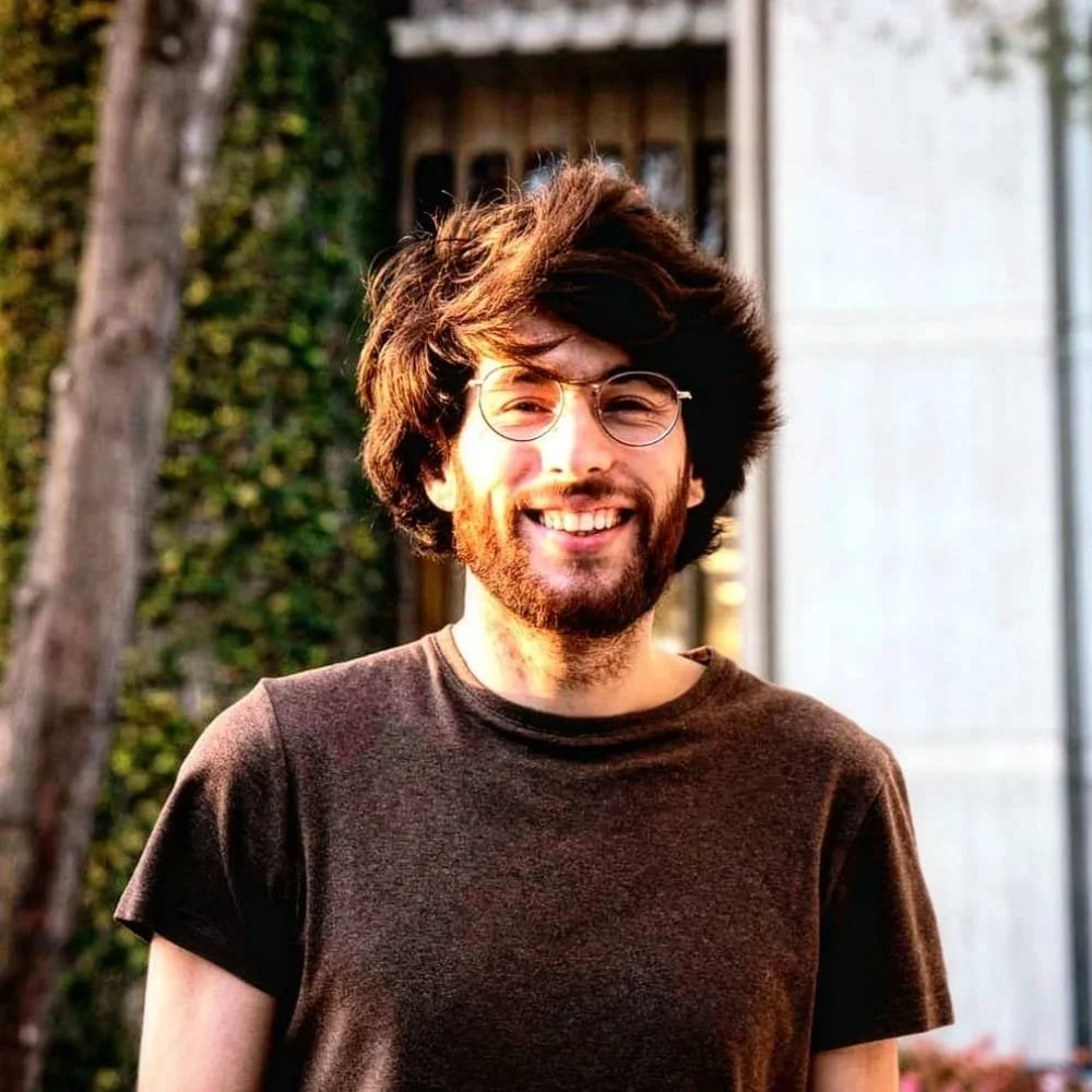 Smiling young man with curly brown hair, beard, glasses, wearing a dark t-shirt, outside in front of a house with trees and ivy.