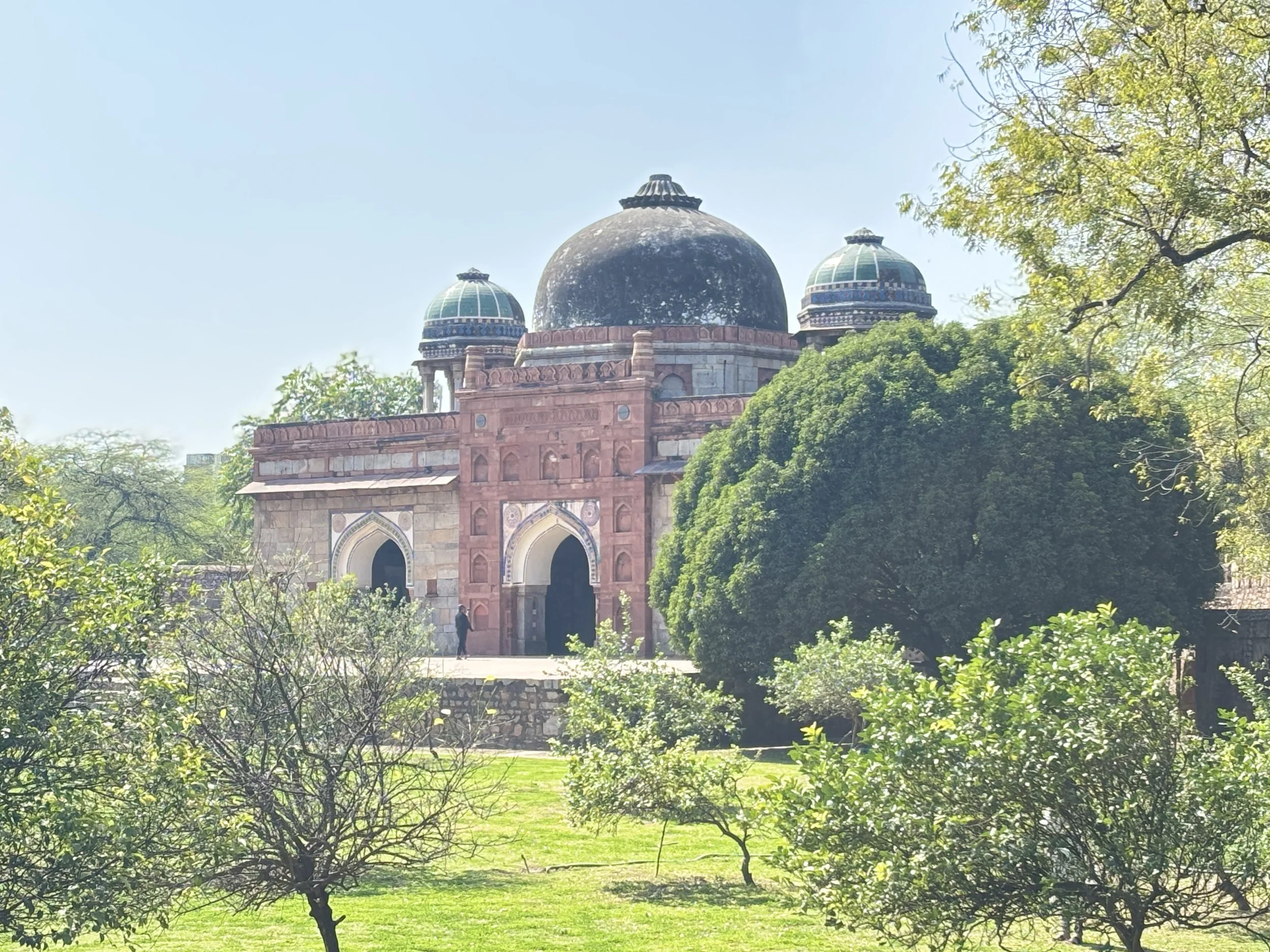 Historical monument with large black central dome and two smaller domed towers on either side, surrounded by greenery and trees, under a clear blue sky.
