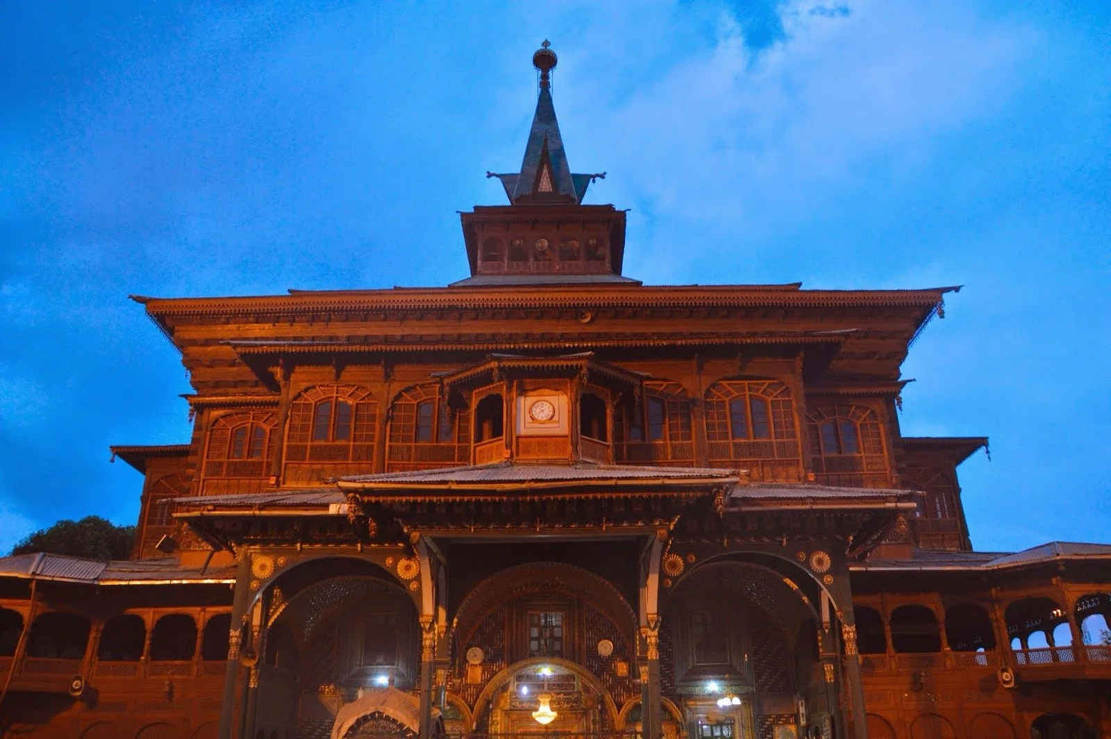 A large, ornate wooden building with multiple levels and a prominent clock on the front, set against a blue evening sky.