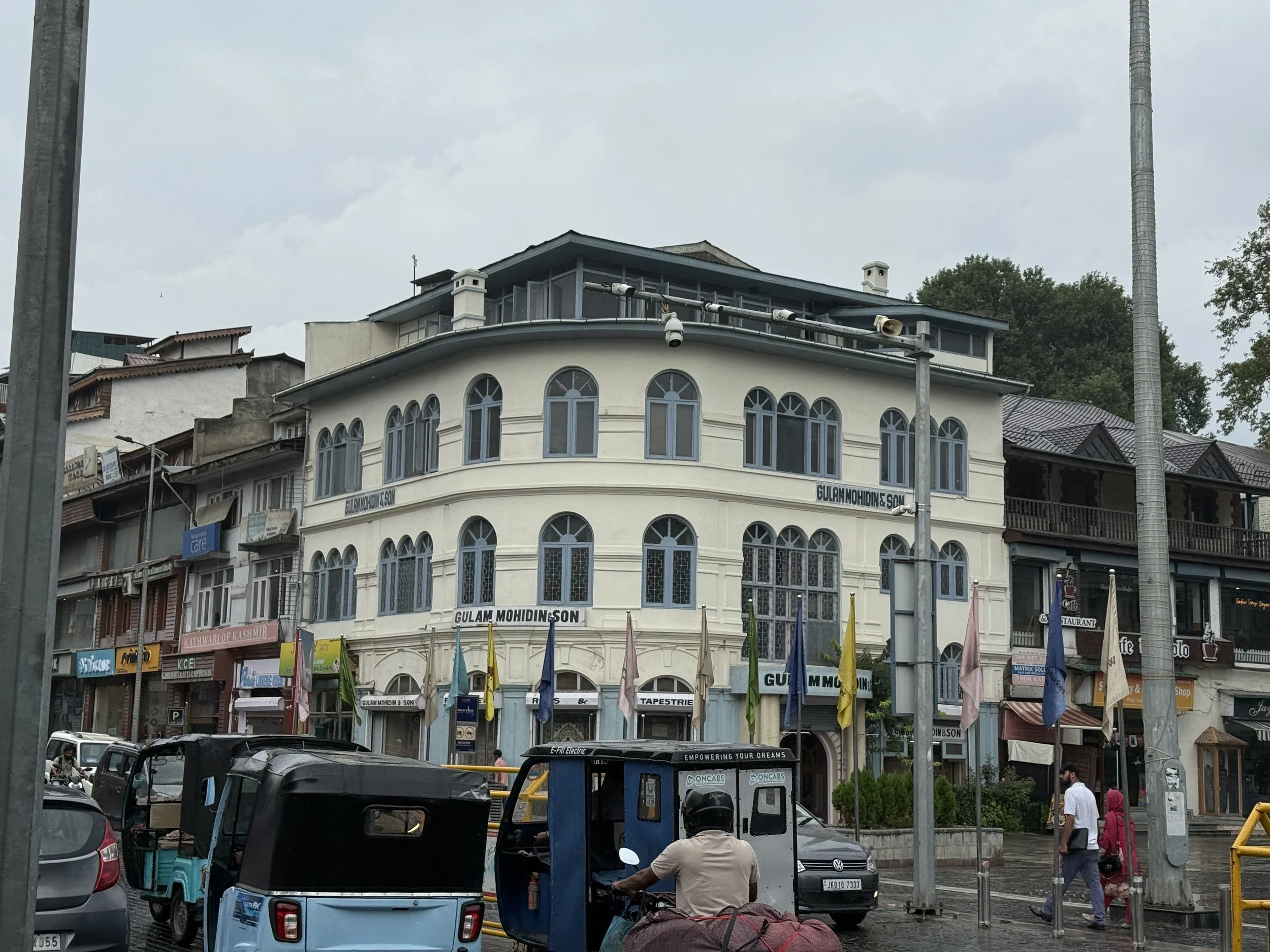 Street view with a white multi-story building, colorful flags, parked vehicles, motorcycles, pedestrians, and street lamps under cloudy sky.