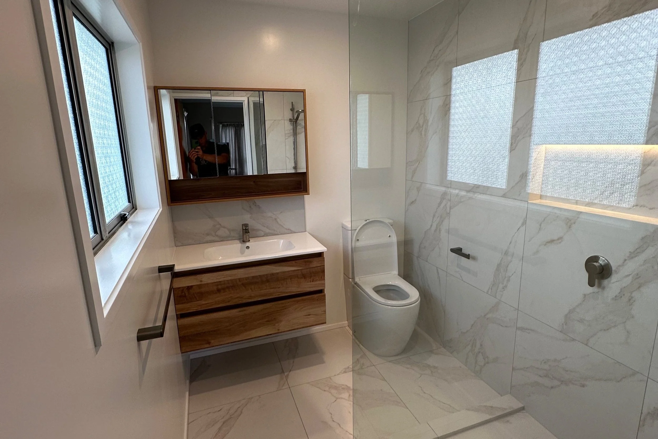 Modern bathroom renovation with wooden vanity, white sink, marble accents, toilet, large mirror, window, and glass shower enclosure.