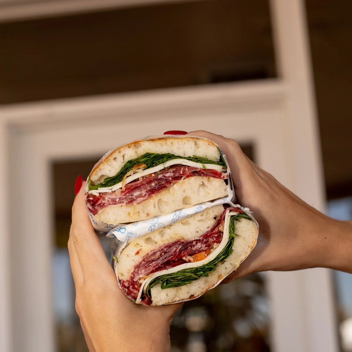 Close-up of a half sandwich with salami, rocket, and cheese held in both hands. Sandwich is called Paulies
