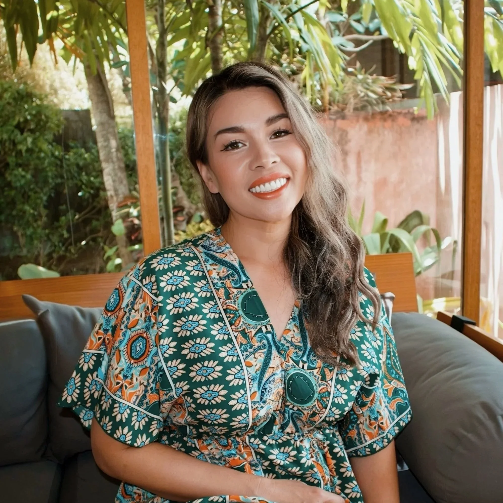 A smiling woman with light brown hair styled in a braid, wearing a colorful patterned shirt, poses on a sandy beach at sunset, with rocks and trees in the background near the ocean.