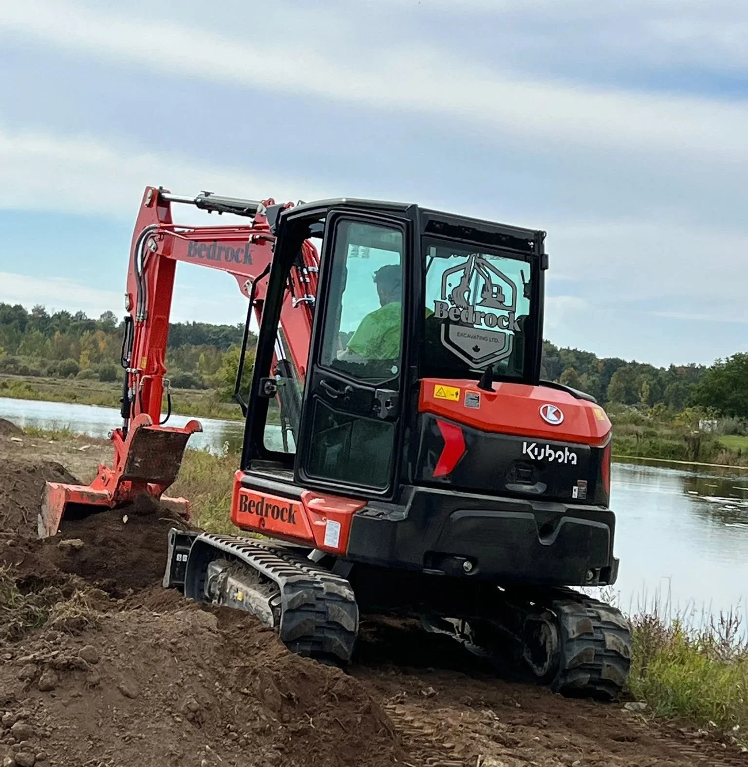 A person operating a red and black Kubota mini excavator labeled 'Bedrock Excavating Ltd.' on a dirt bank near a body of water, with trees and hills in the background.
