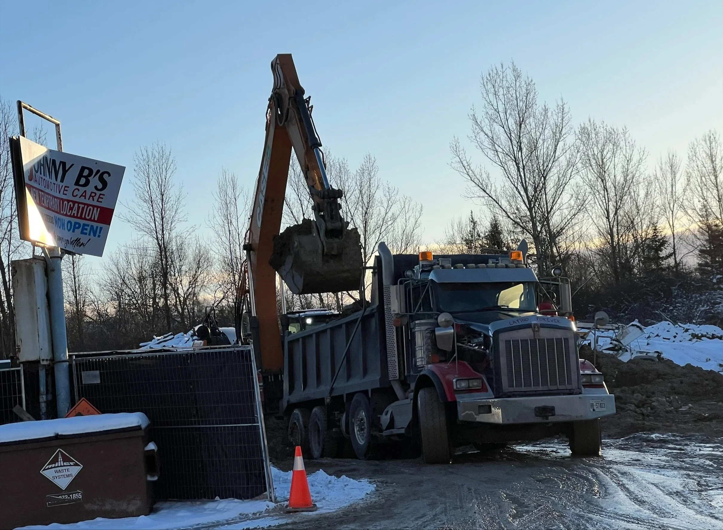 Construction site with a large dump truck and an excavator lifting a load of dirt, surrounded by snow and trees.