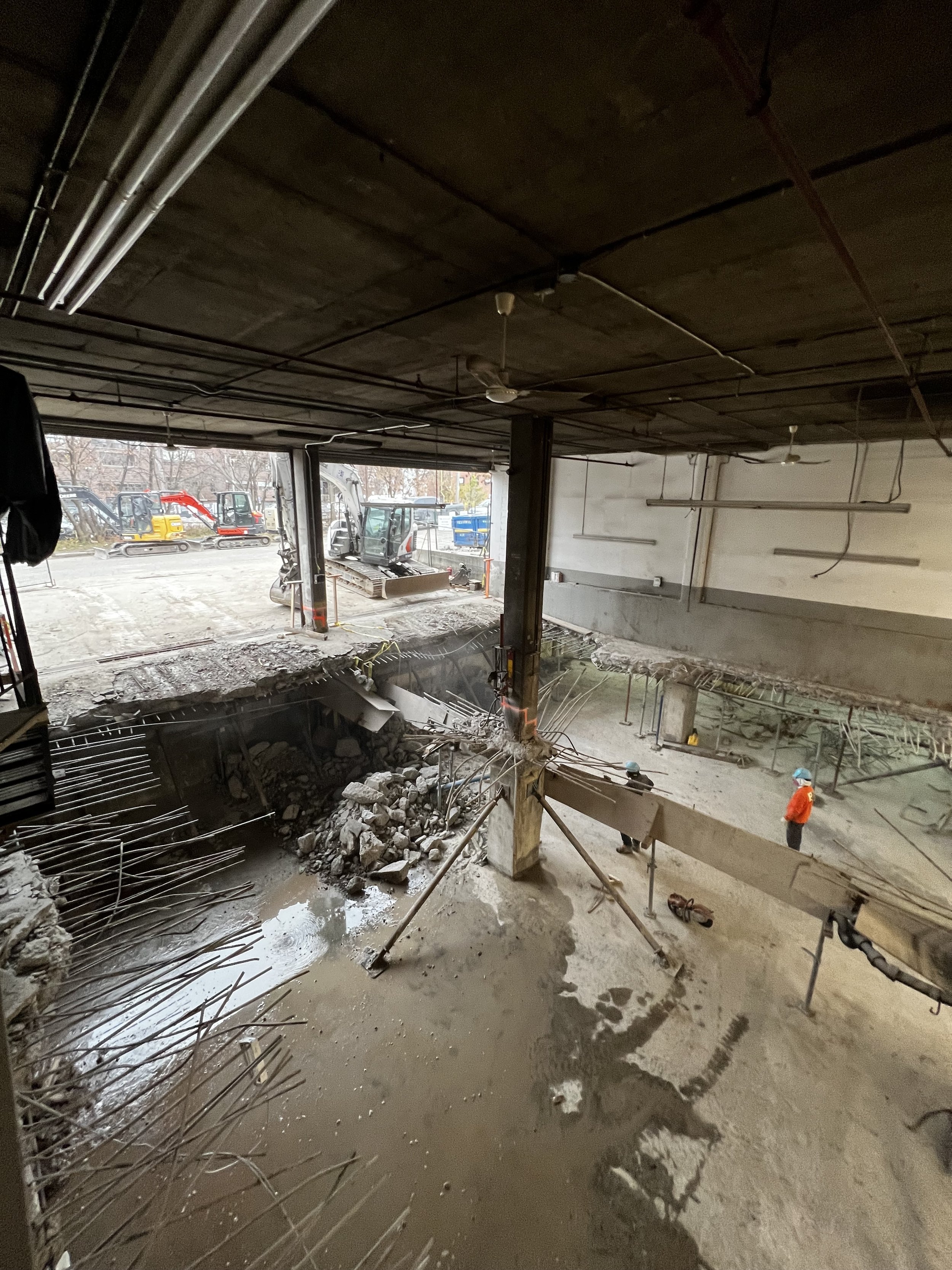 Construction site with exposed concrete, steel rebar, construction workers, and machinery seen through an open garage area.