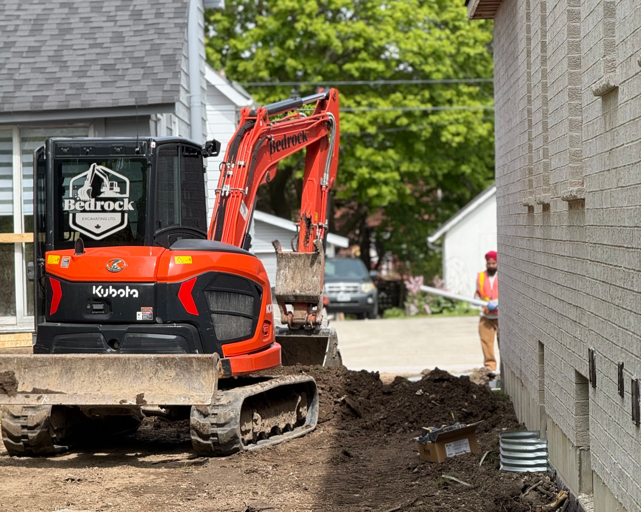 Construction site with an orange mini excavator labeled 'Kubota' and 'Bedrock' parked on dirt. In the background, a worker wearing a yellow safety vest and red hat is visible.