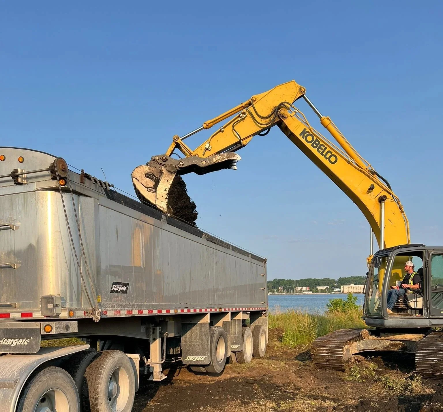 A yellow Kobelco excavator is loading dark soil or dirt into a large silver dump truck near a body of water with buildings in the background, under a clear blue sky.