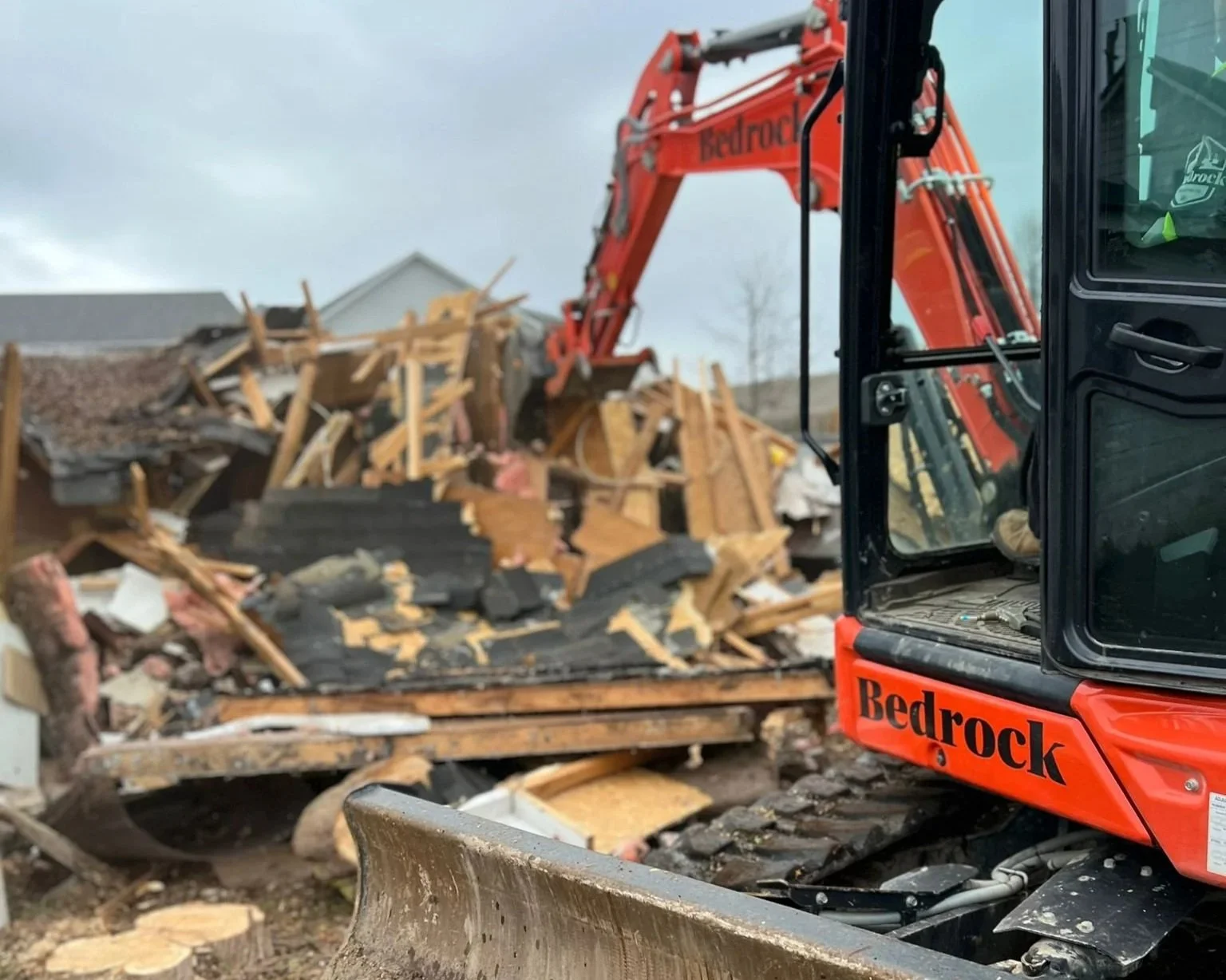 A red Bedrock excavator tearing down a wooden structure with debris scattered around on a cloudy day.